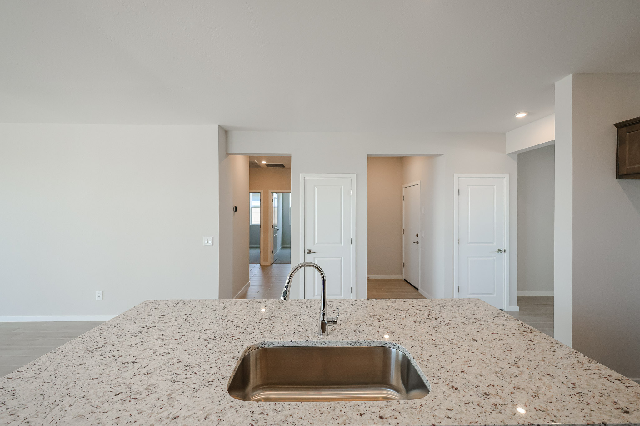 View from a kitchen island with a sink facing a hallway and three closed white doors in a modern, unfurnished home with neutral walls and floors.