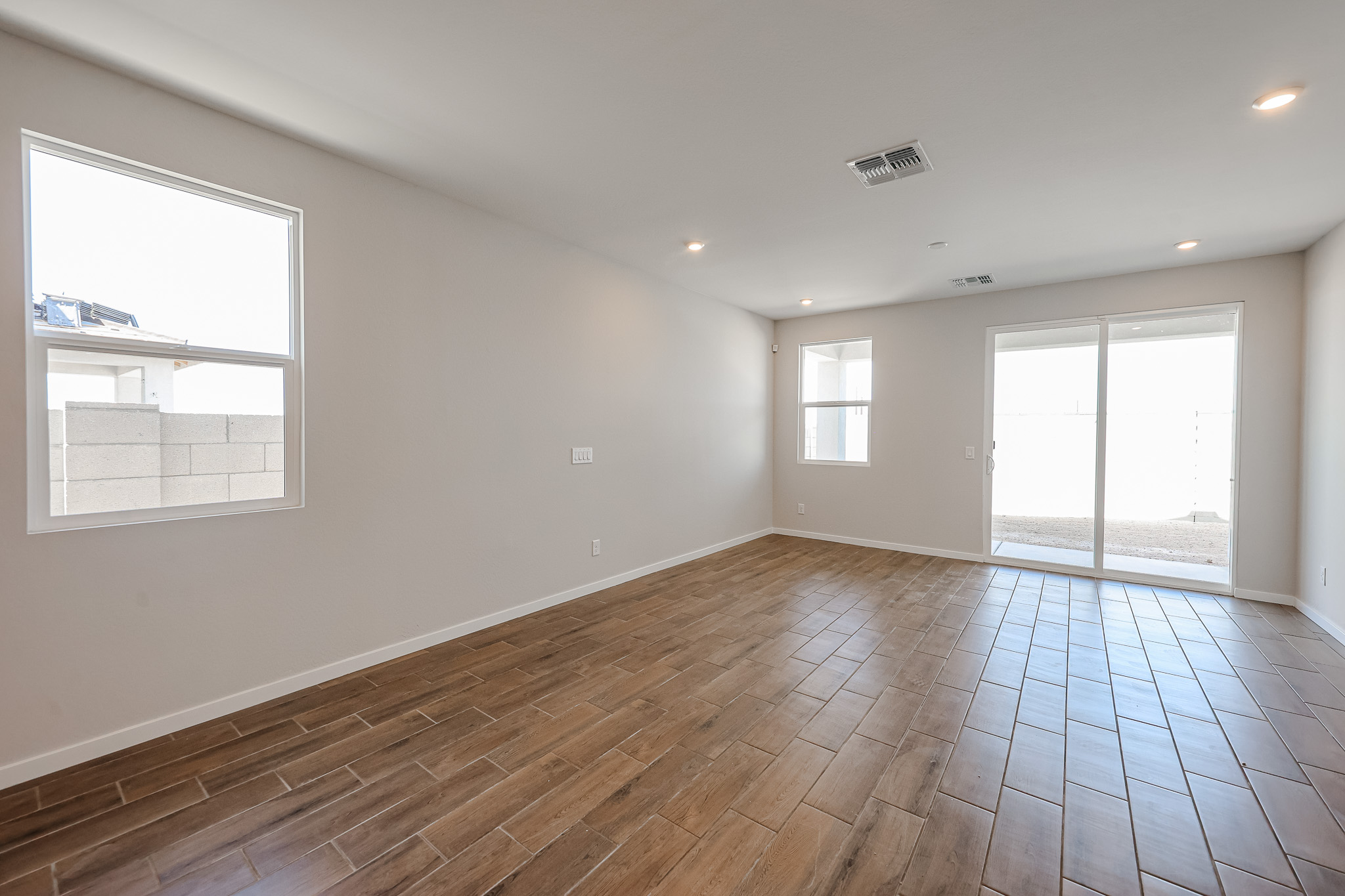 Empty room with light brown wood-look tile flooring, white walls, recessed ceiling lights, two windows, and a sliding glass door leading to an outdoor area.