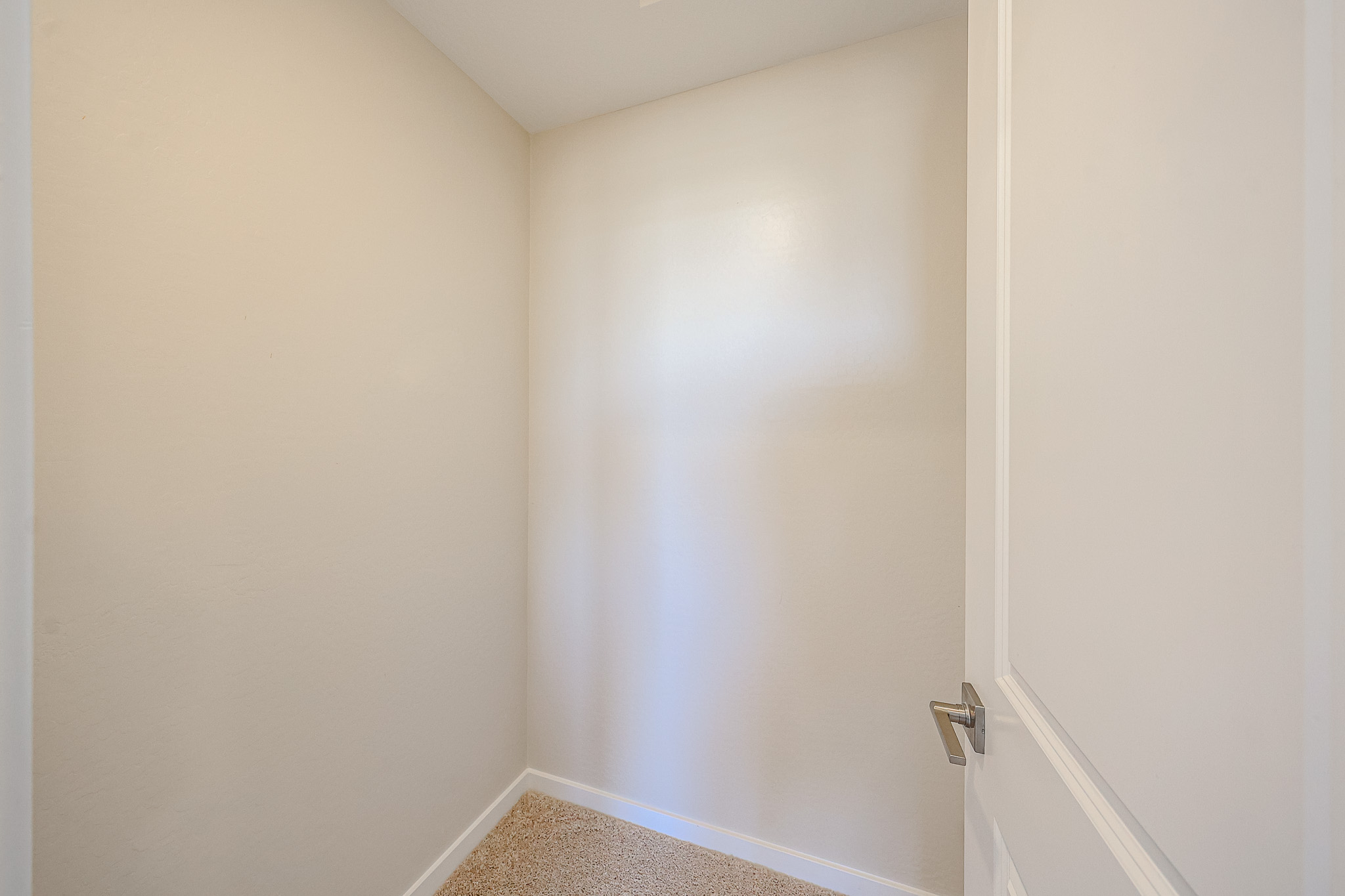 Small, empty beige room with carpet flooring, white trim, and a partially open white door. Walls and ceiling are plain with no decorations or fixtures visible.