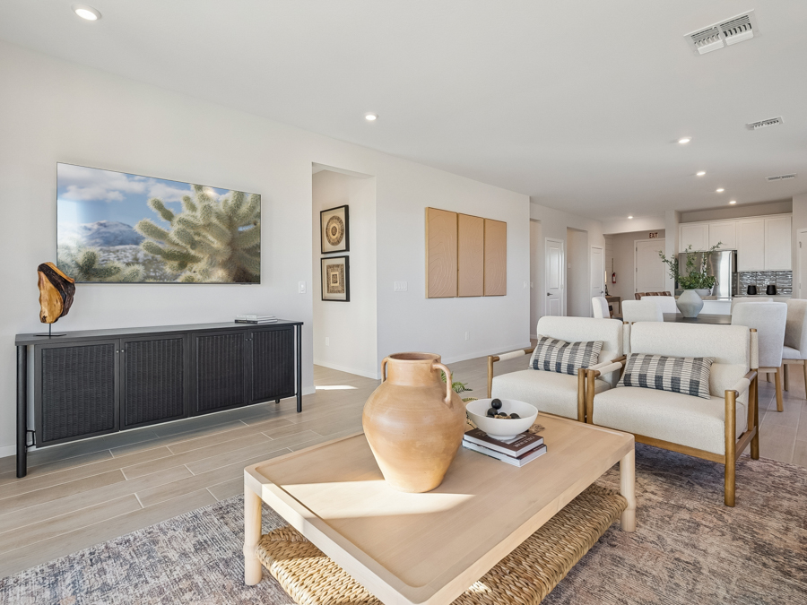 Modern living room with a TV, two armchairs, a coffee table with decor, and a view into the dining area and kitchen; neutral tones dominate the space.