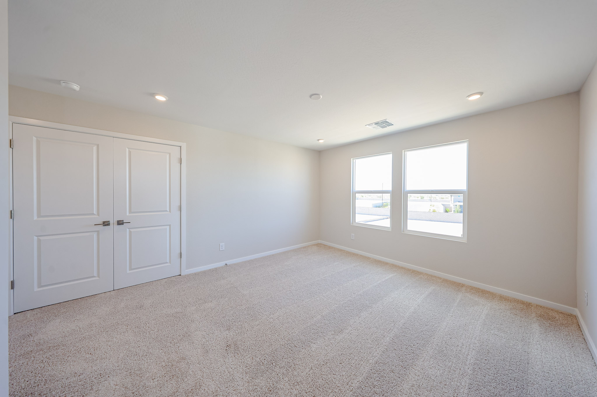 Empty, carpeted bedroom with white walls, double doors on the left, and two windows on the right letting in natural light.