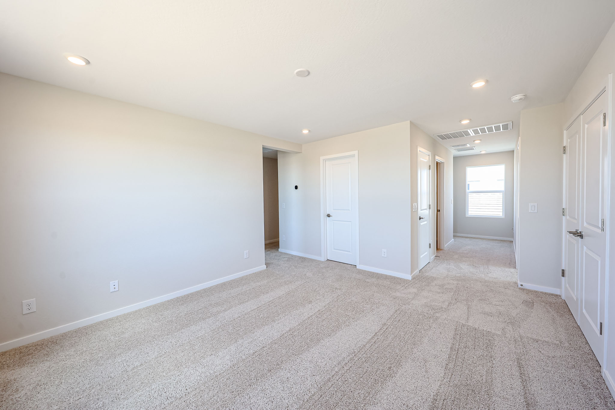 Empty, carpeted room with beige walls, recessed ceiling lights, two closed white doors, and a hallway leading to a window at the far end.