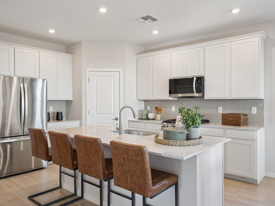 Modern kitchen with white cabinets, stainless steel appliances, an island with sink, brown leather barstools, and potted plants on the counter. Bright, clean, and well-lit space.