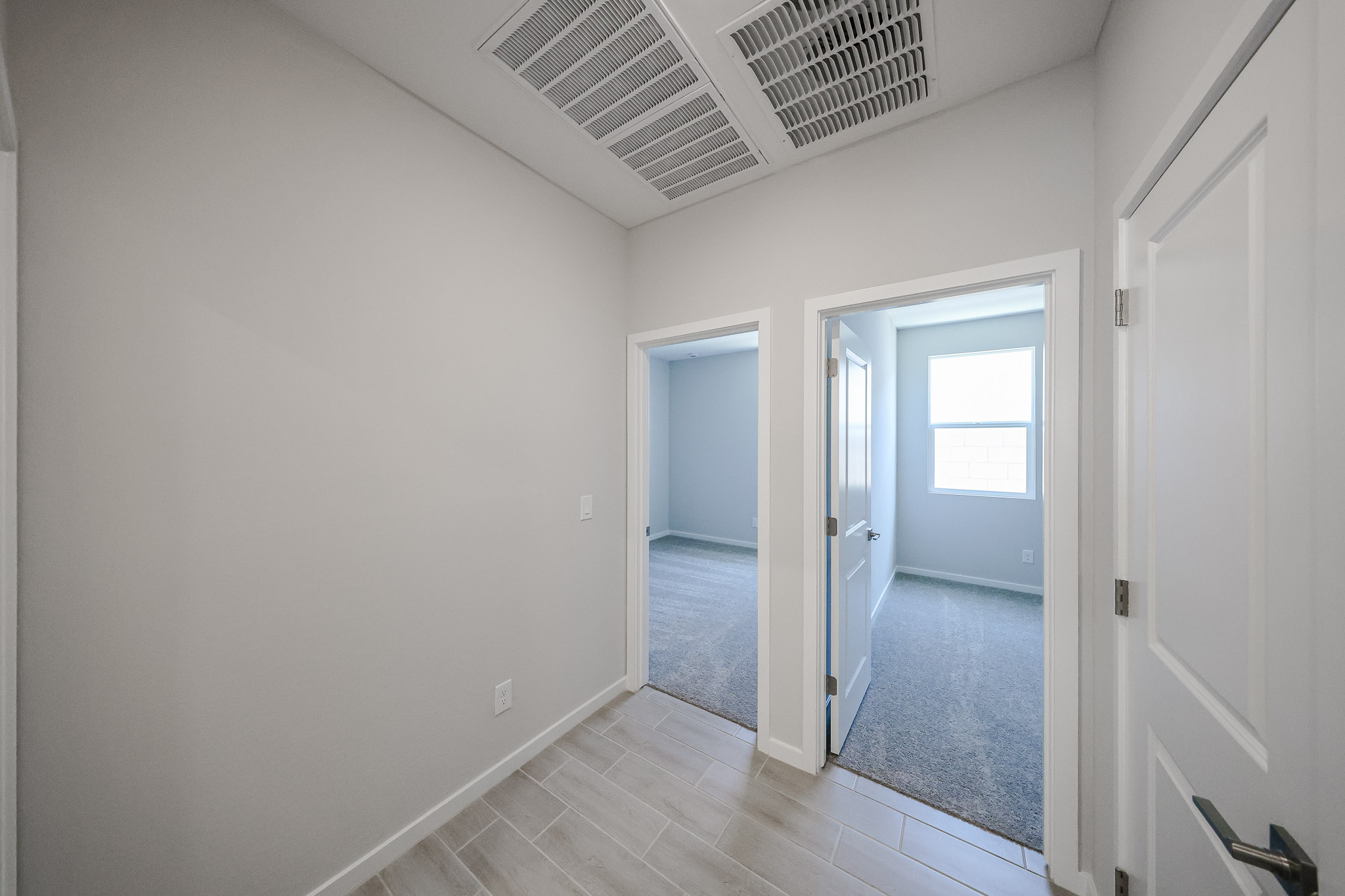 A hallway with light tile flooring and two open doors leading to carpeted rooms, white walls, and air vents on the ceiling.