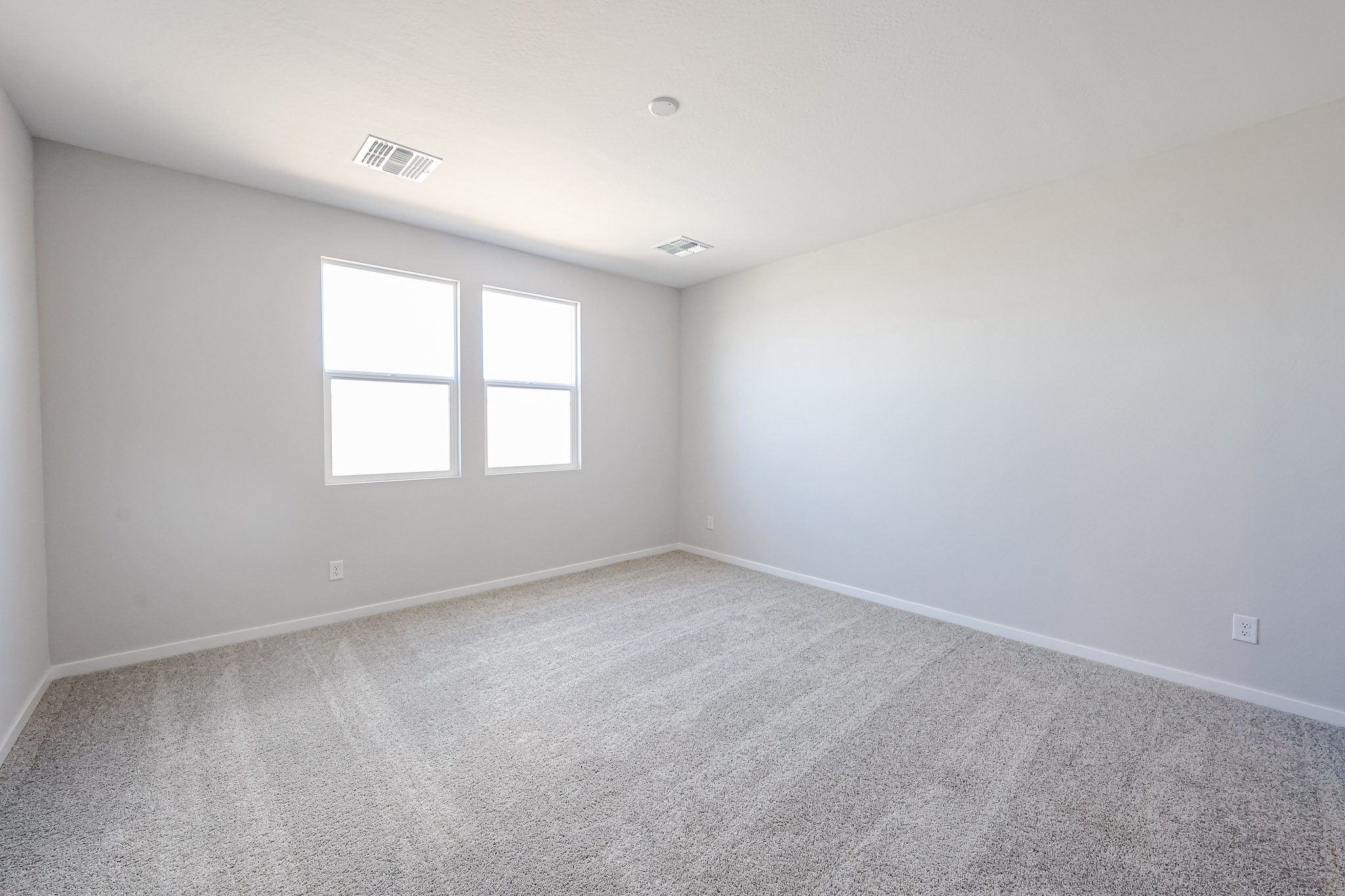 Empty room with light gray carpet, white walls, two windows, and ceiling vents. Natural light fills the space.