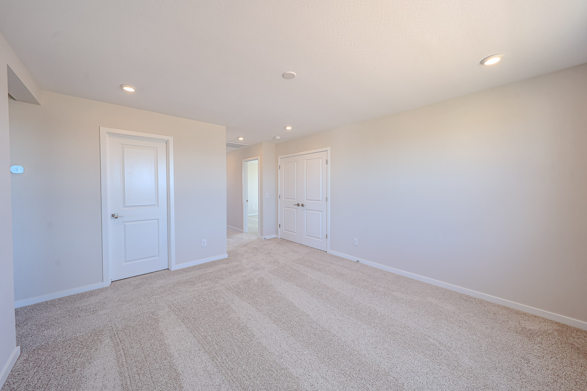 Empty bedroom with beige carpet, light-colored walls, recessed ceiling lights, and three closed white doors.