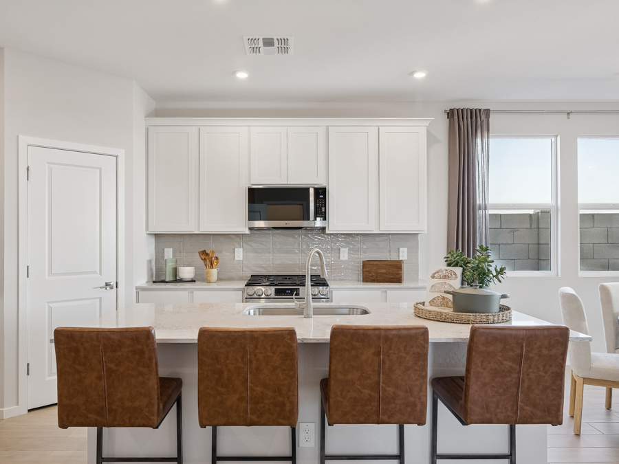 Modern kitchen with white cabinets, stainless steel appliances, a center island with sink, and four brown leather bar stools. Dining table and large windows are visible in the background.