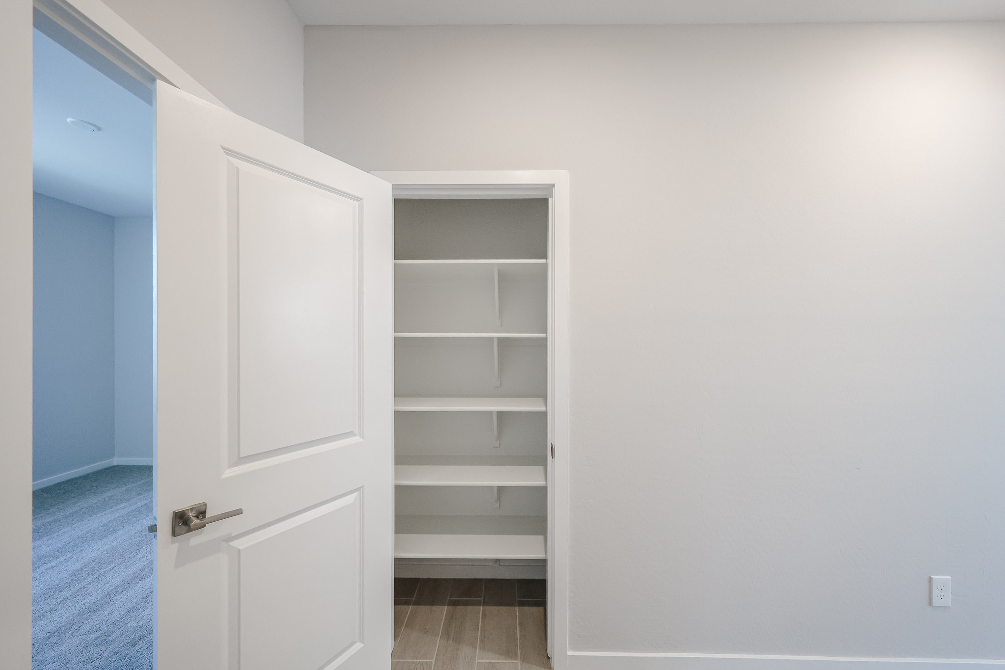Open white door reveals a small closet with four empty white shelves, set against light-colored walls and a tiled floor.