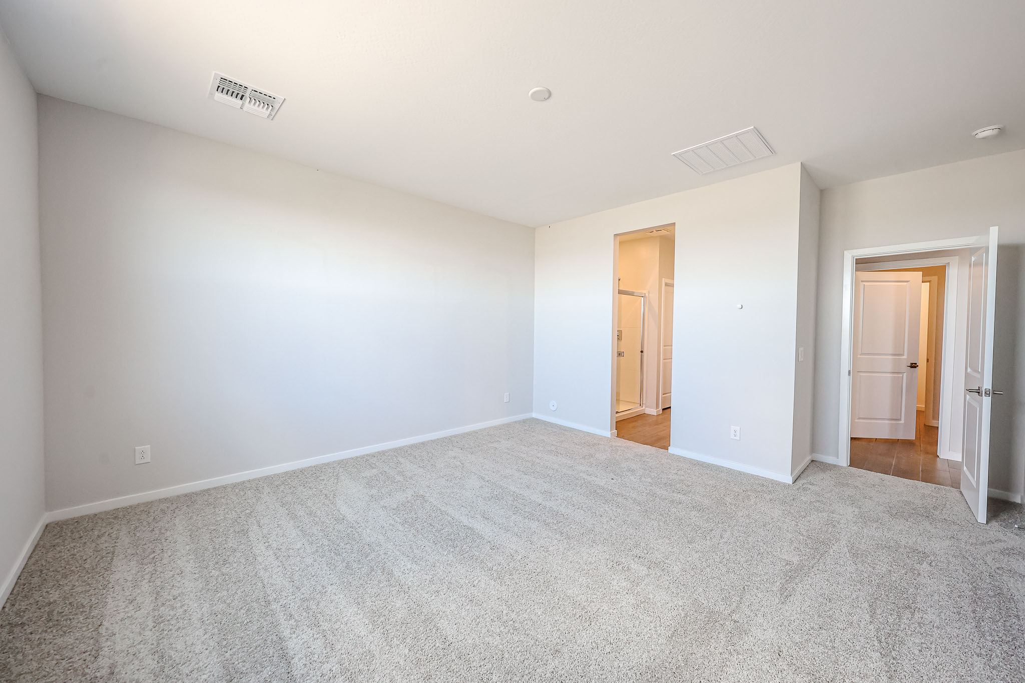 Empty carpeted room with white walls, two open doors—one leading to a bathroom, the other to a hallway. Ceiling vent and light fixtures are visible.