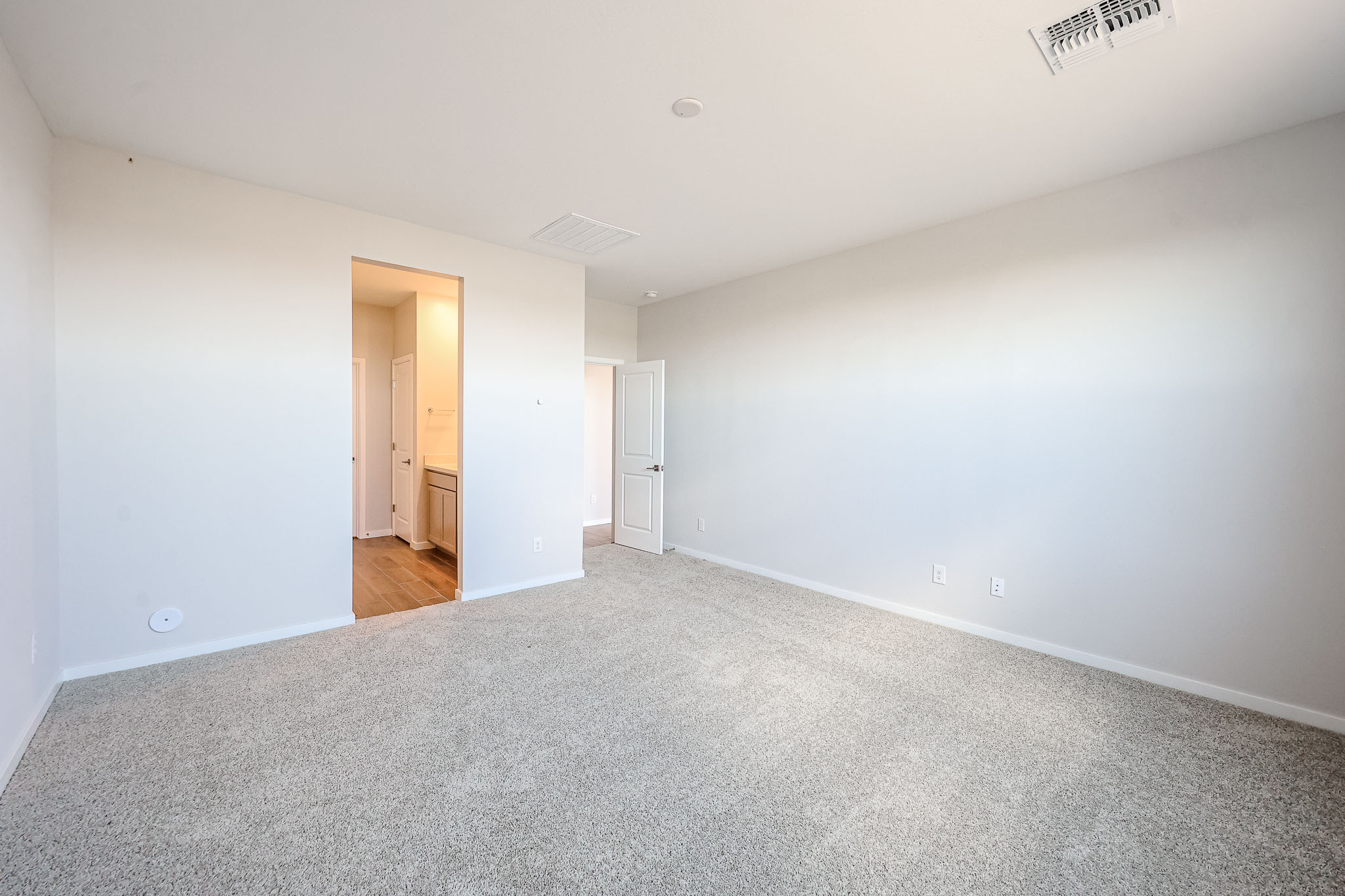 Empty room with light-colored carpet, white walls, and two open doorways leading to a hallway and bathroom. Ceiling vent and electrical outlets are visible.
