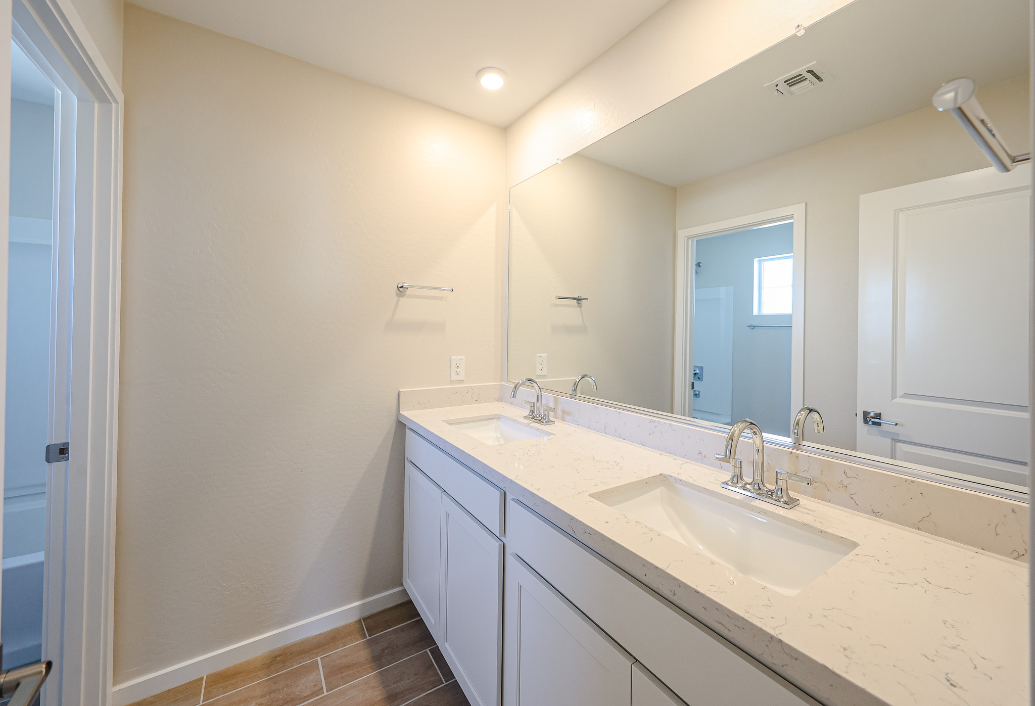 Modern bathroom with double sinks, white cabinets, a large wall mirror, tan walls, and brown tile flooring. A towel rack and electrical outlets are visible. Room is brightly lit.