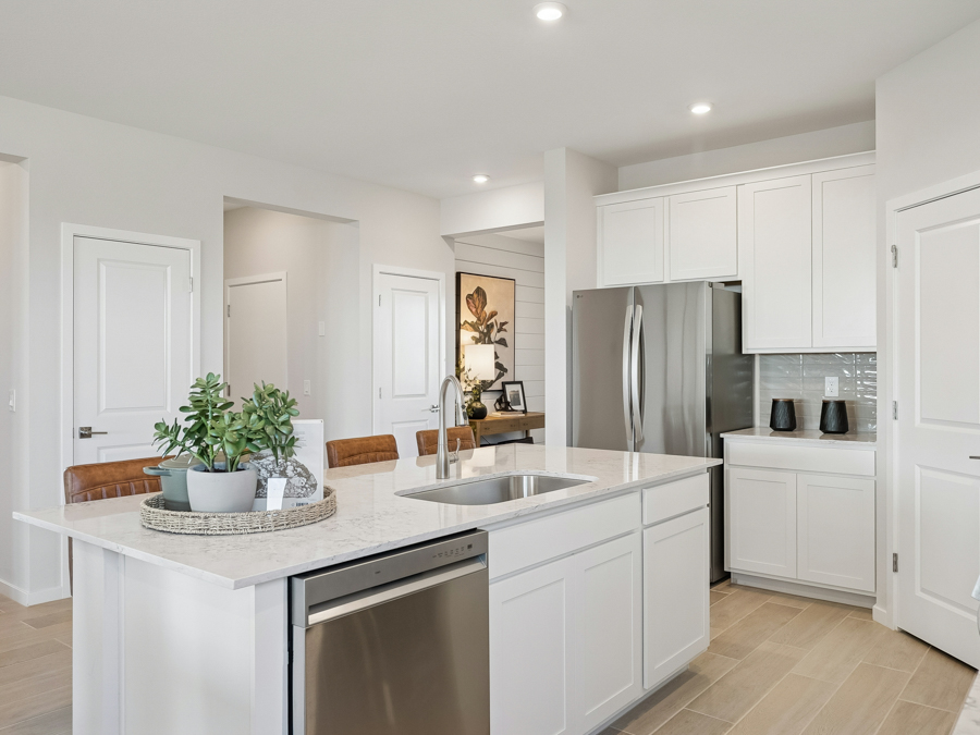 Modern kitchen with white cabinets, stainless steel appliances, a large island with sink, and a tray of potted plants. Two brown chairs are positioned at the island.