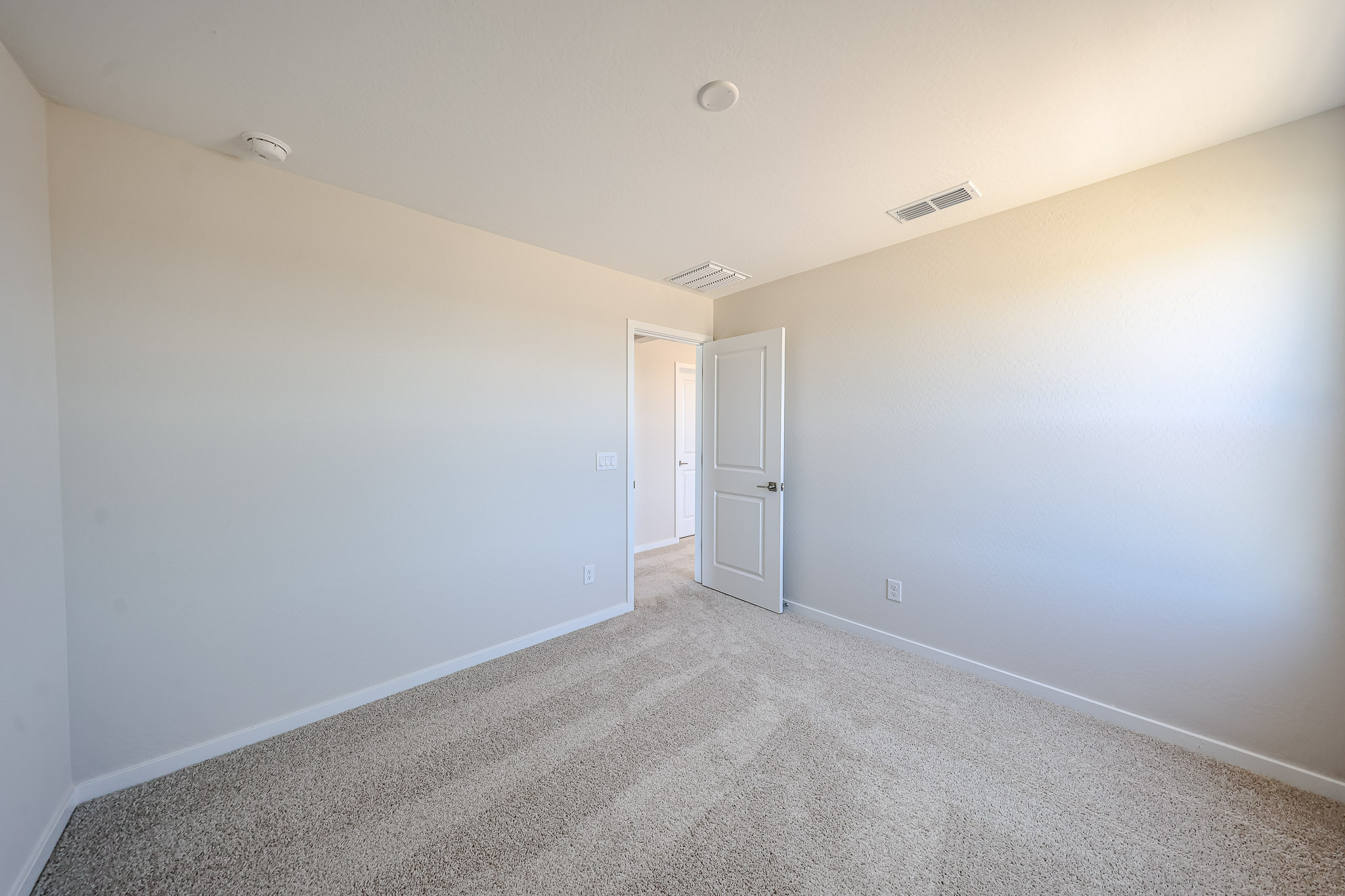 Empty room with beige carpet, cream-colored walls, a white door slightly open, ceiling vent, and bright natural light from an unseen window.