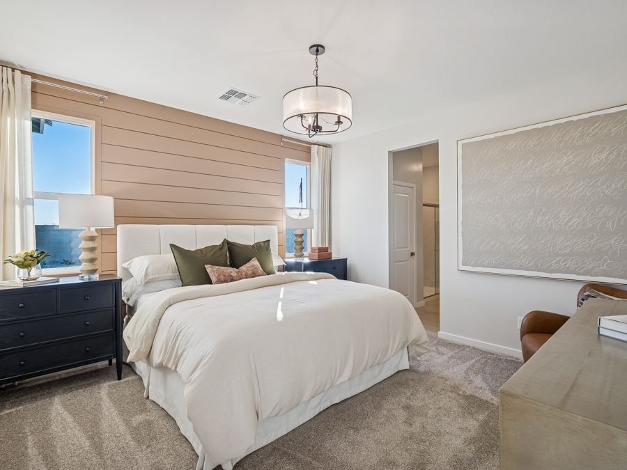 Modern bedroom with a beige bed, two dark nightstands, a large abstract artwork, and a ceiling light fixture. Carpeting covers the floor, and natural light enters through two windows.