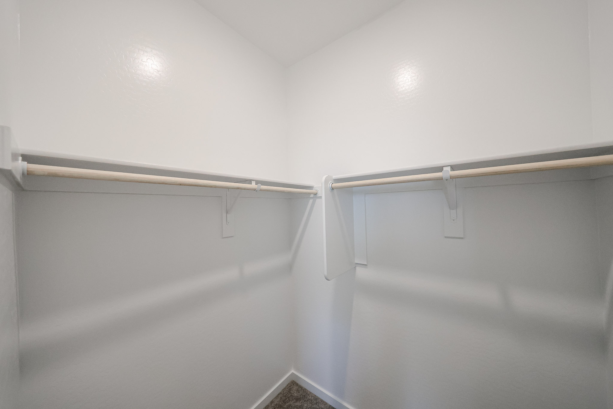 Empty walk-in closet with white walls, two wooden hanging rods, and carpeted flooring. The closet is well-lit and clean.
