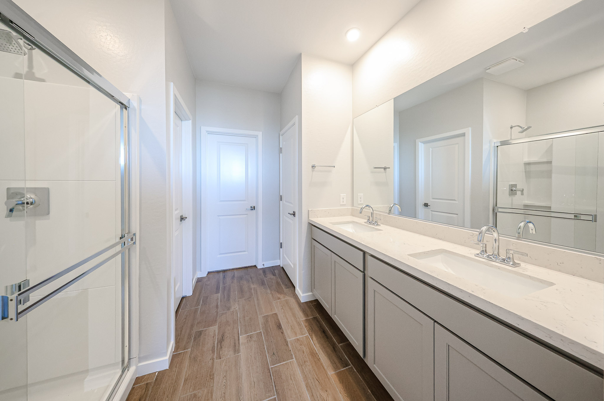 Modern bathroom with a double vanity, large mirror, glass shower enclosure, and wood-look tile flooring. Walls and cabinetry are light-colored, giving the room a bright and clean appearance.
