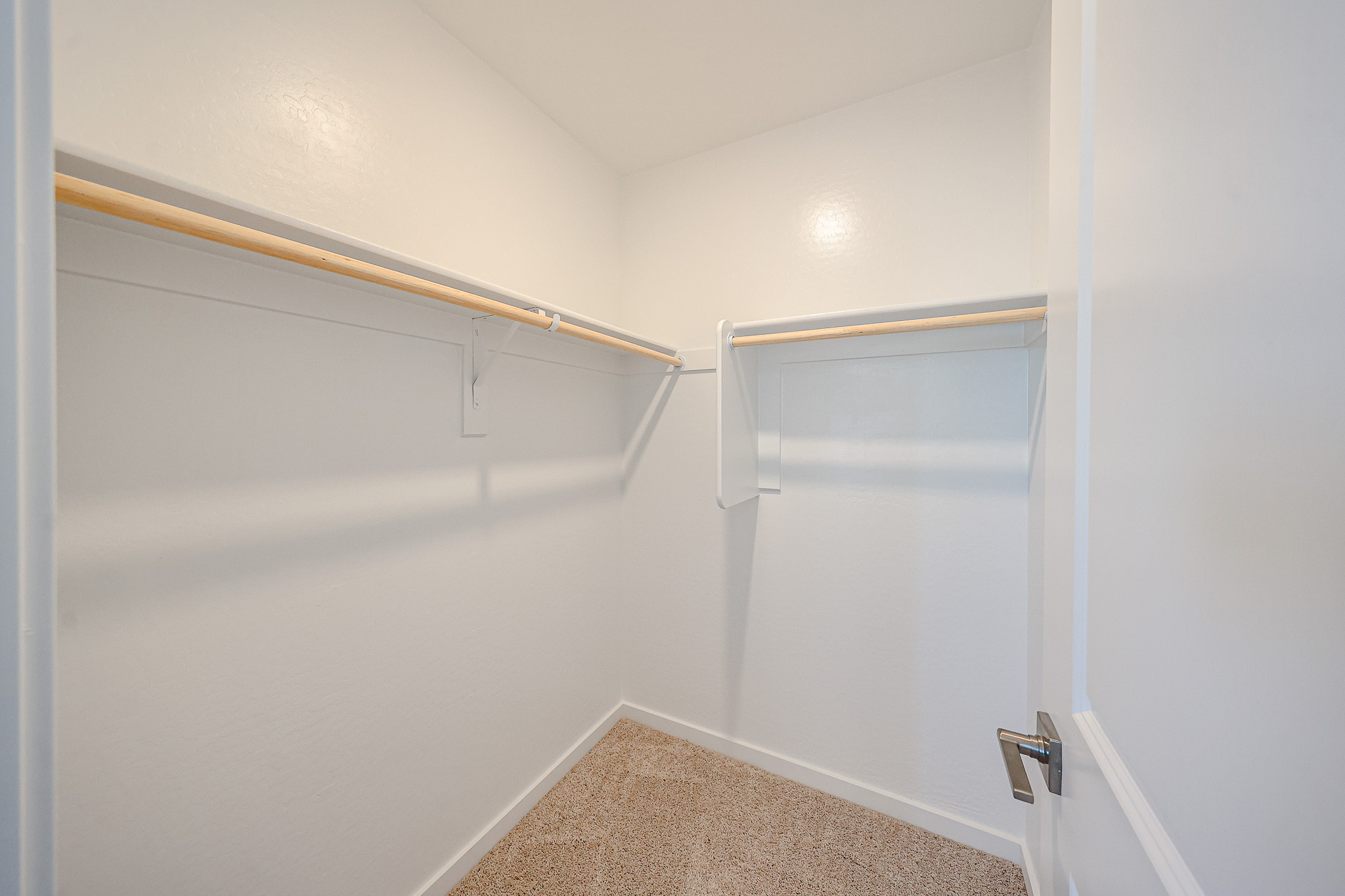 Empty walk-in closet with white walls, beige carpet, and wooden hanging rods on two sides.