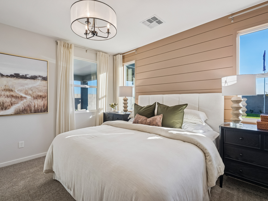 Modern bedroom with a beige accent wall behind a bed with white linens, olive green pillows, two bedside tables with lamps, large windows, and a landscape artwork on the left wall.