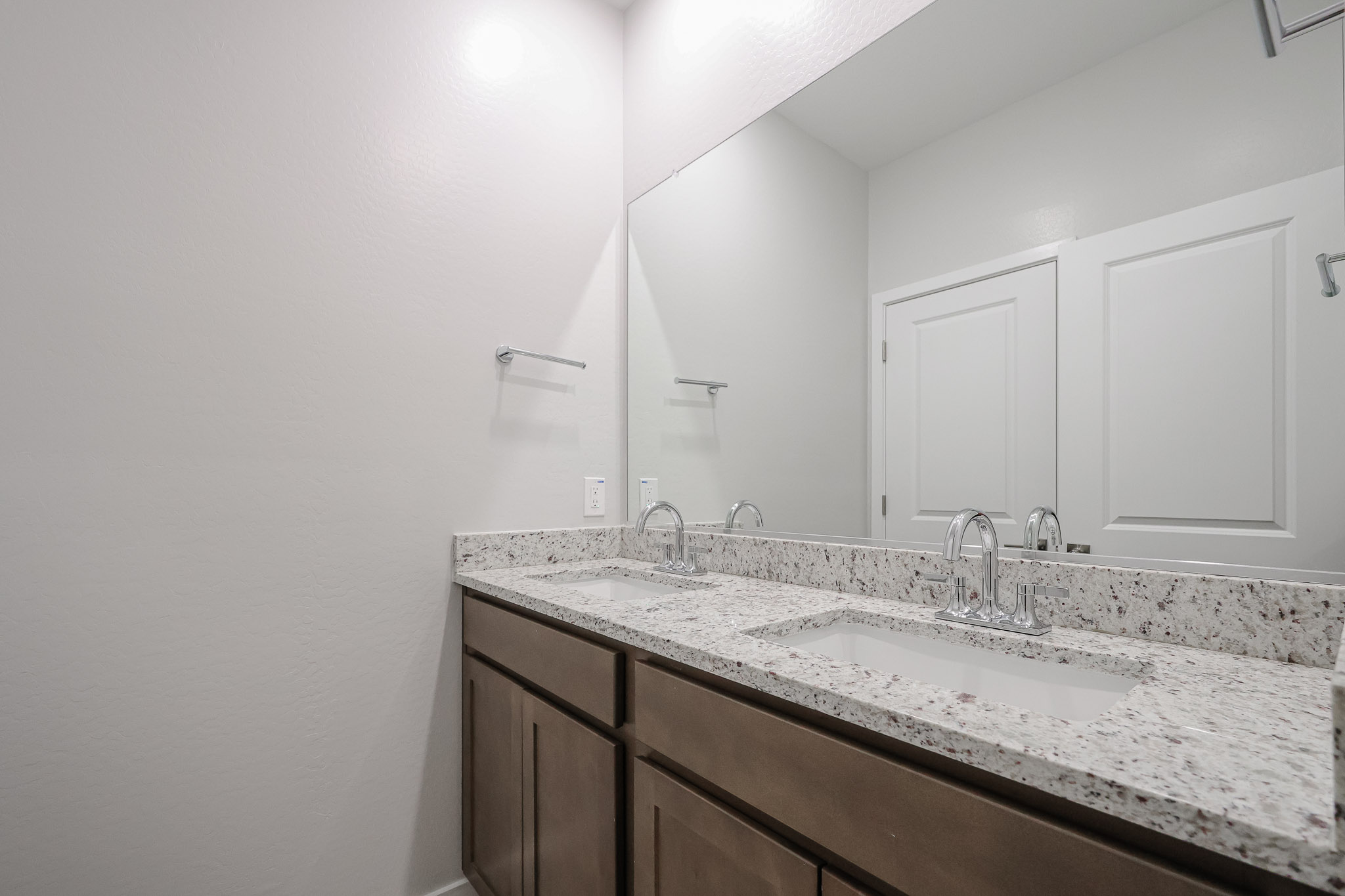Modern bathroom with double sinks, granite countertop, brown cabinets, white walls, a large mirror, and two doors reflected in the mirror.