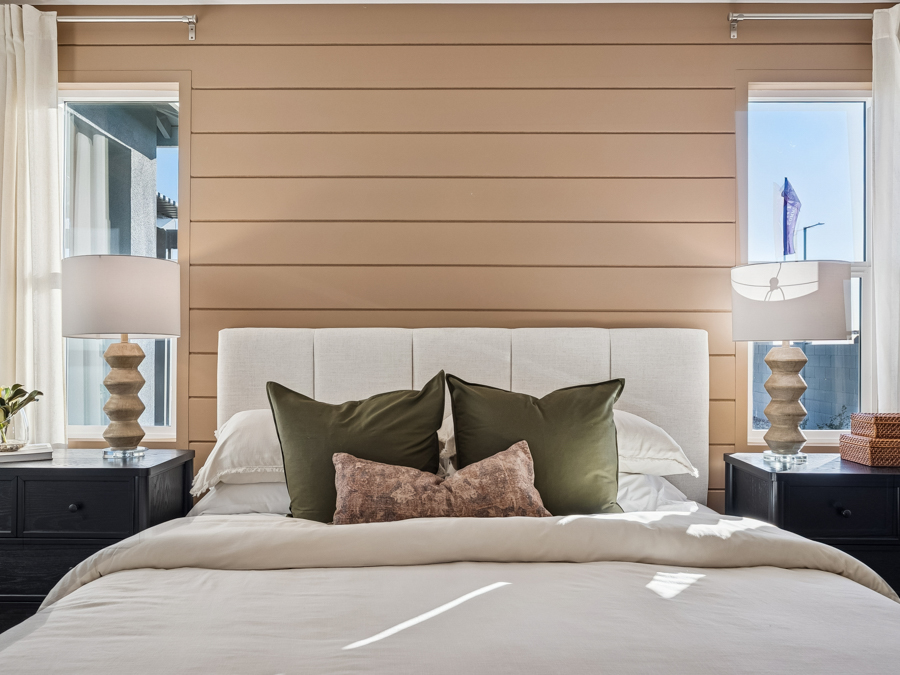 Modern bedroom with a beige shiplap wall, two windows, a bed with green and brown pillows, white bedding, black nightstands, and matching lamps on each side.