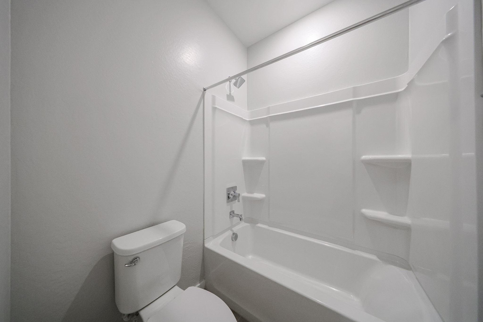 White bathroom with a toilet and a built-in bathtub-shower combo, featuring a showerhead, faucet, and three shelves in a bright, minimal space.