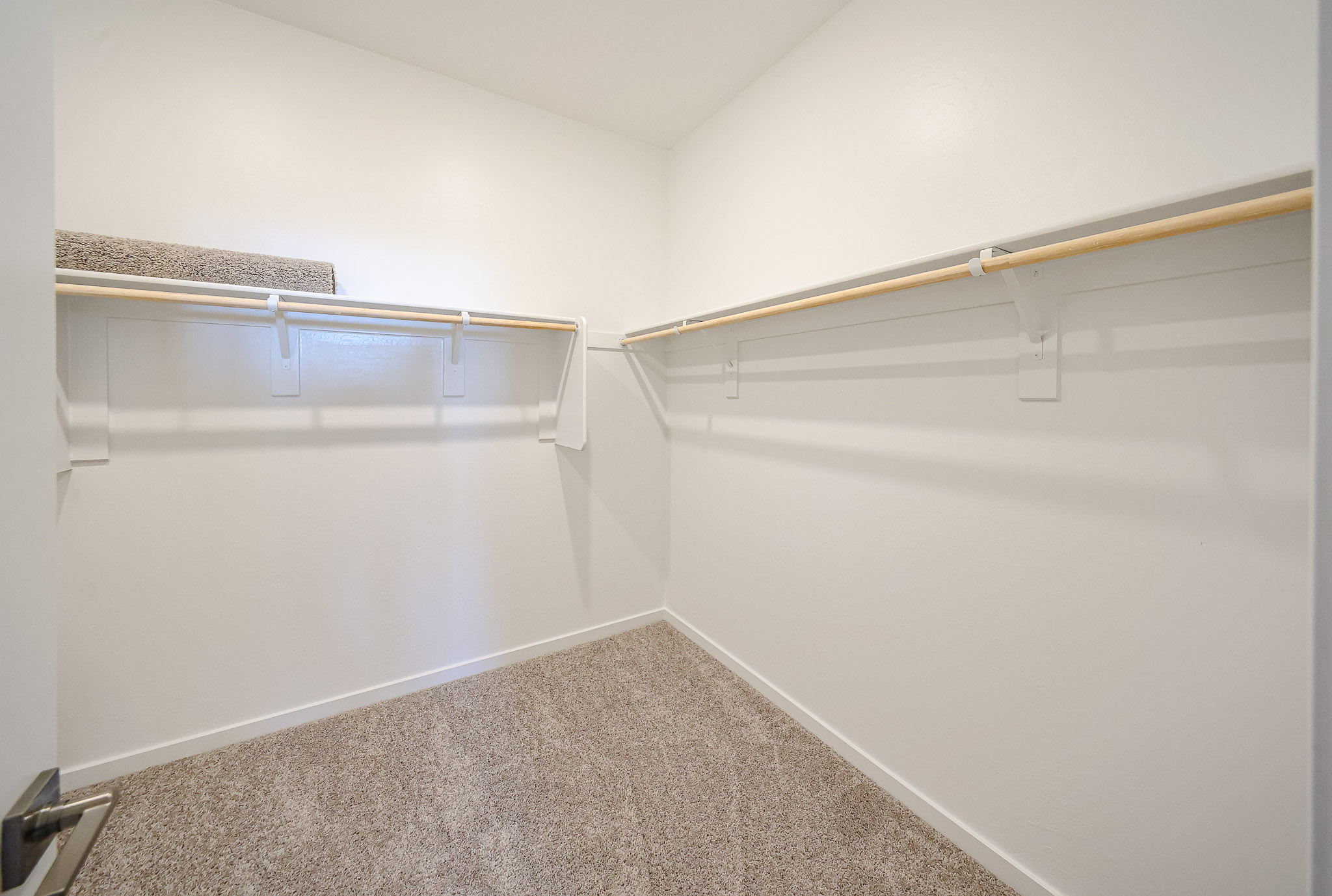 Empty walk-in closet with beige carpet, white walls, wooden rod shelves on each side, and a rolled-up rug on the left shelf.