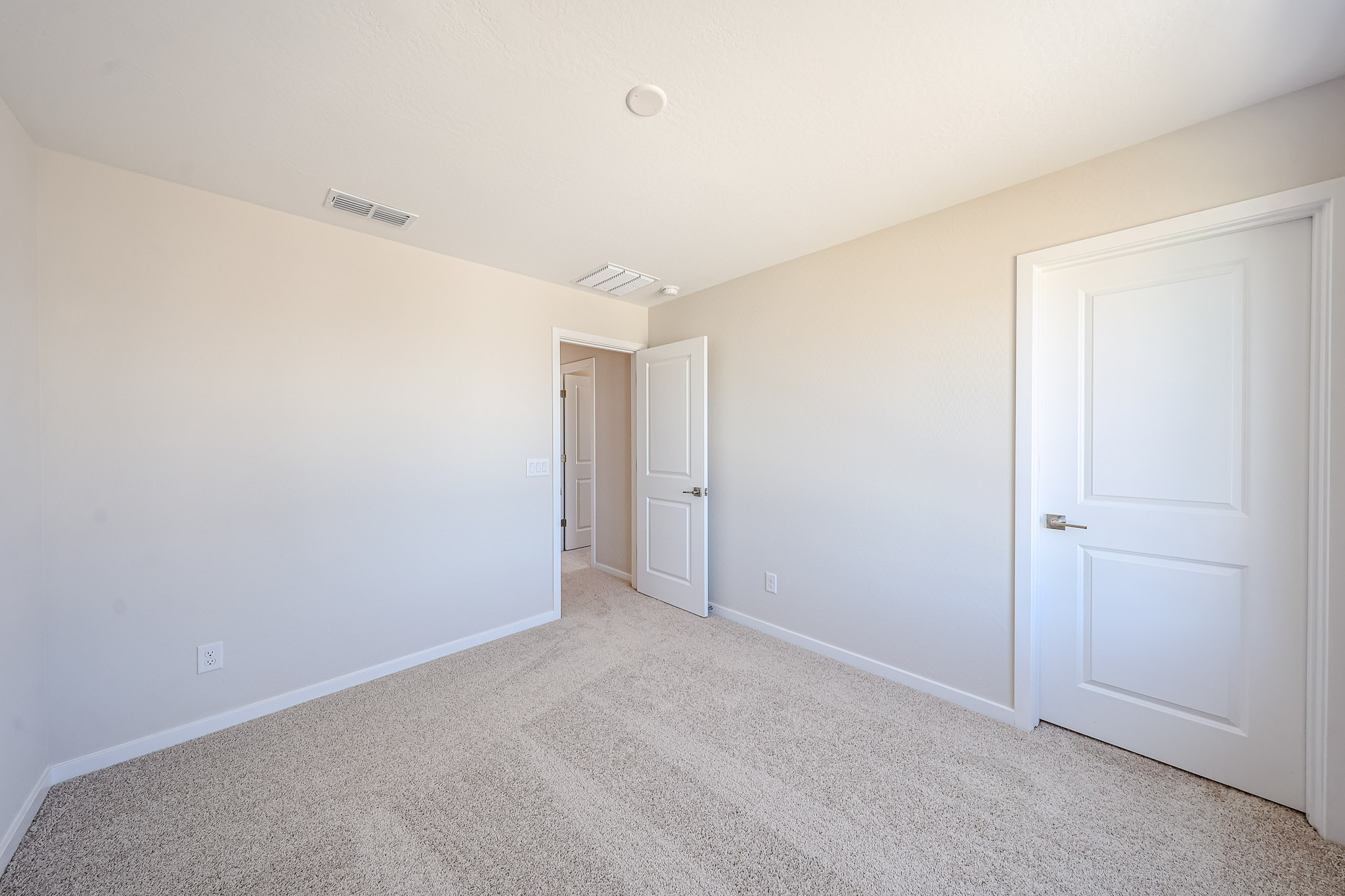 Empty room with beige carpet, off-white walls, and two closed white doors. One door leads to a hallway. Ceiling vent and light fixture also visible.
