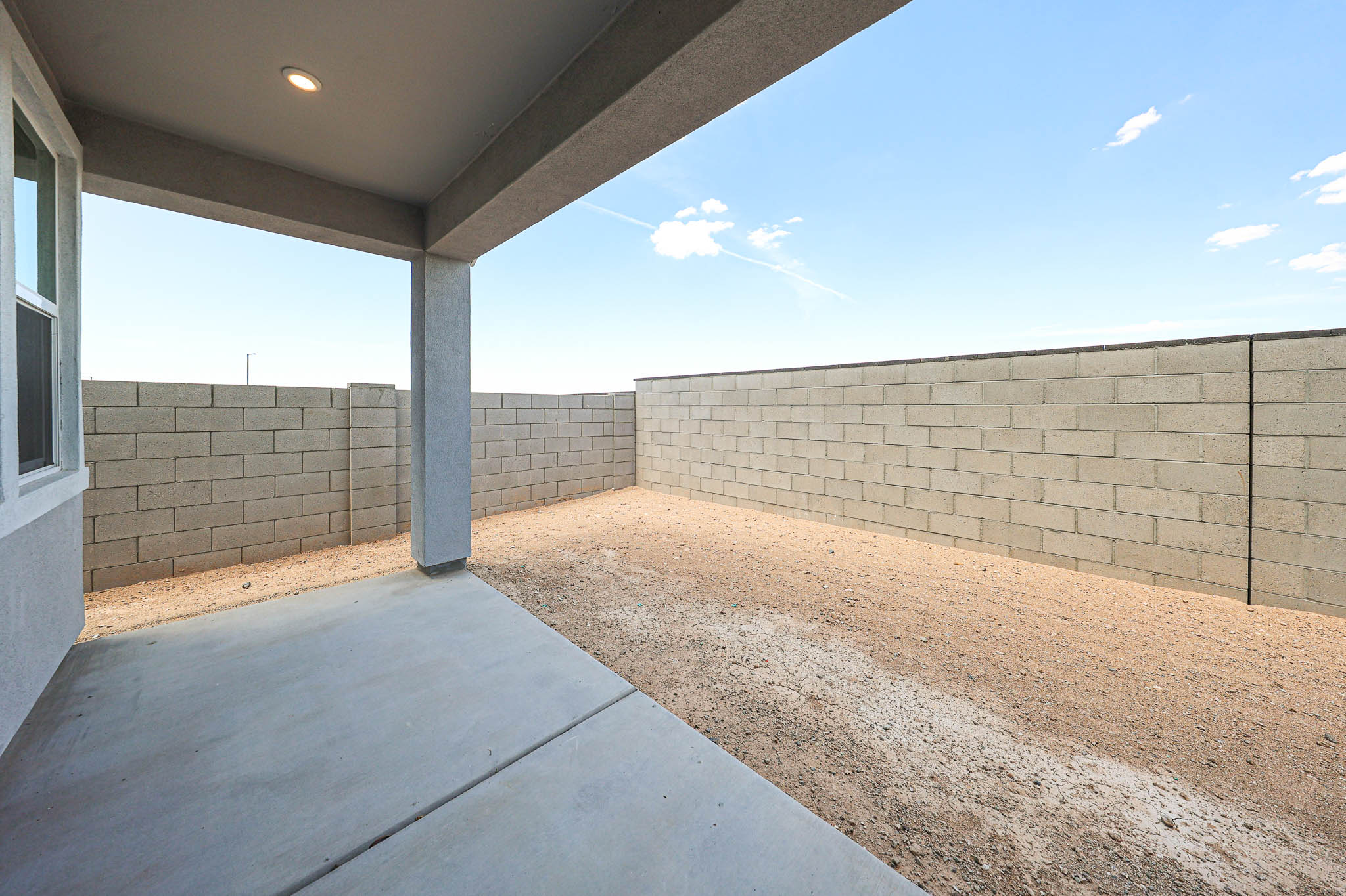 Covered concrete patio with ceiling light, overlooking a small, fenced backyard enclosed by a high cinder block wall under a clear blue sky.