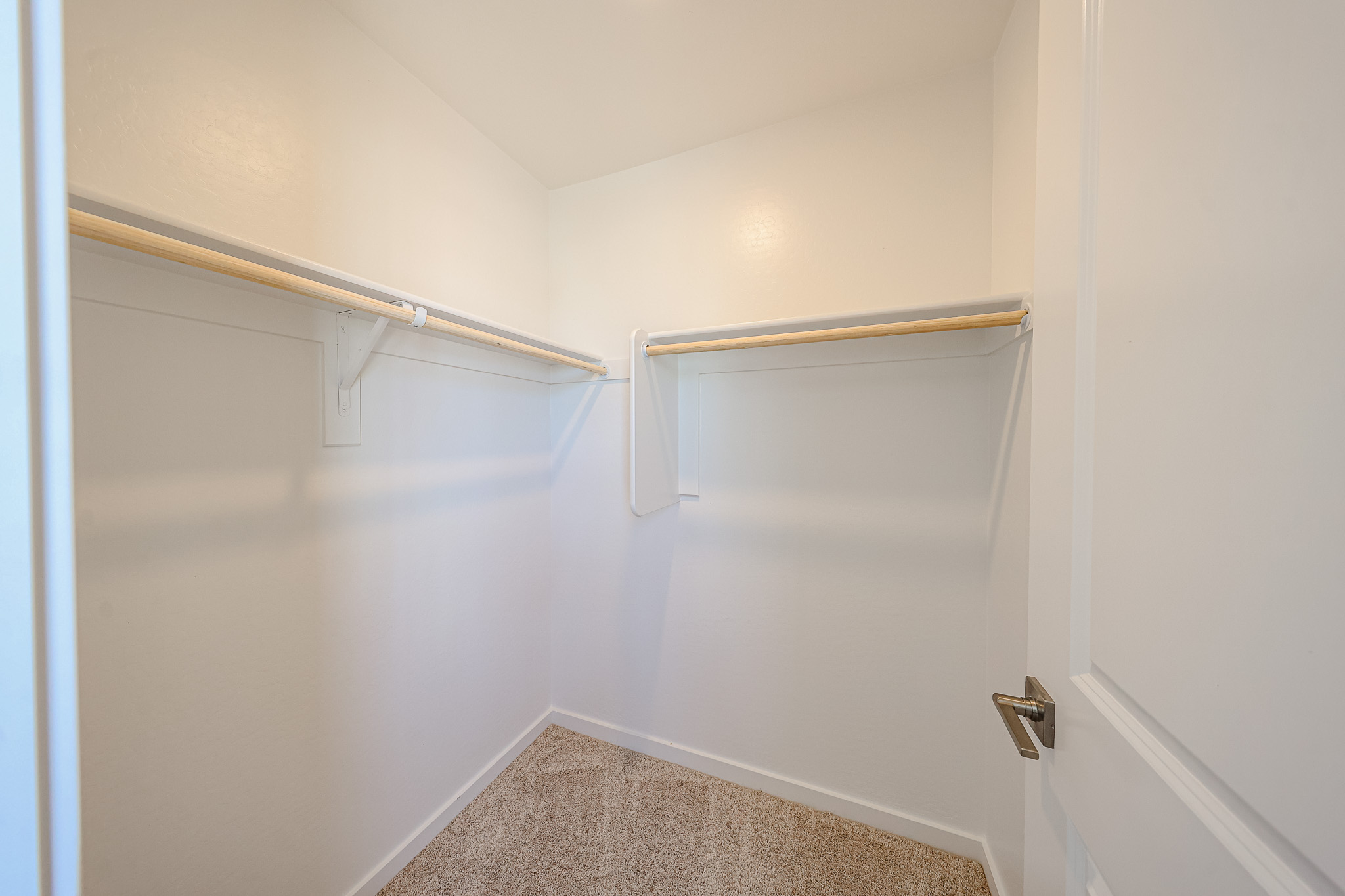 Empty walk-in closet with beige carpet, white walls, and wooden hanging rods on two sides. The door is open, providing a view of the interior space.