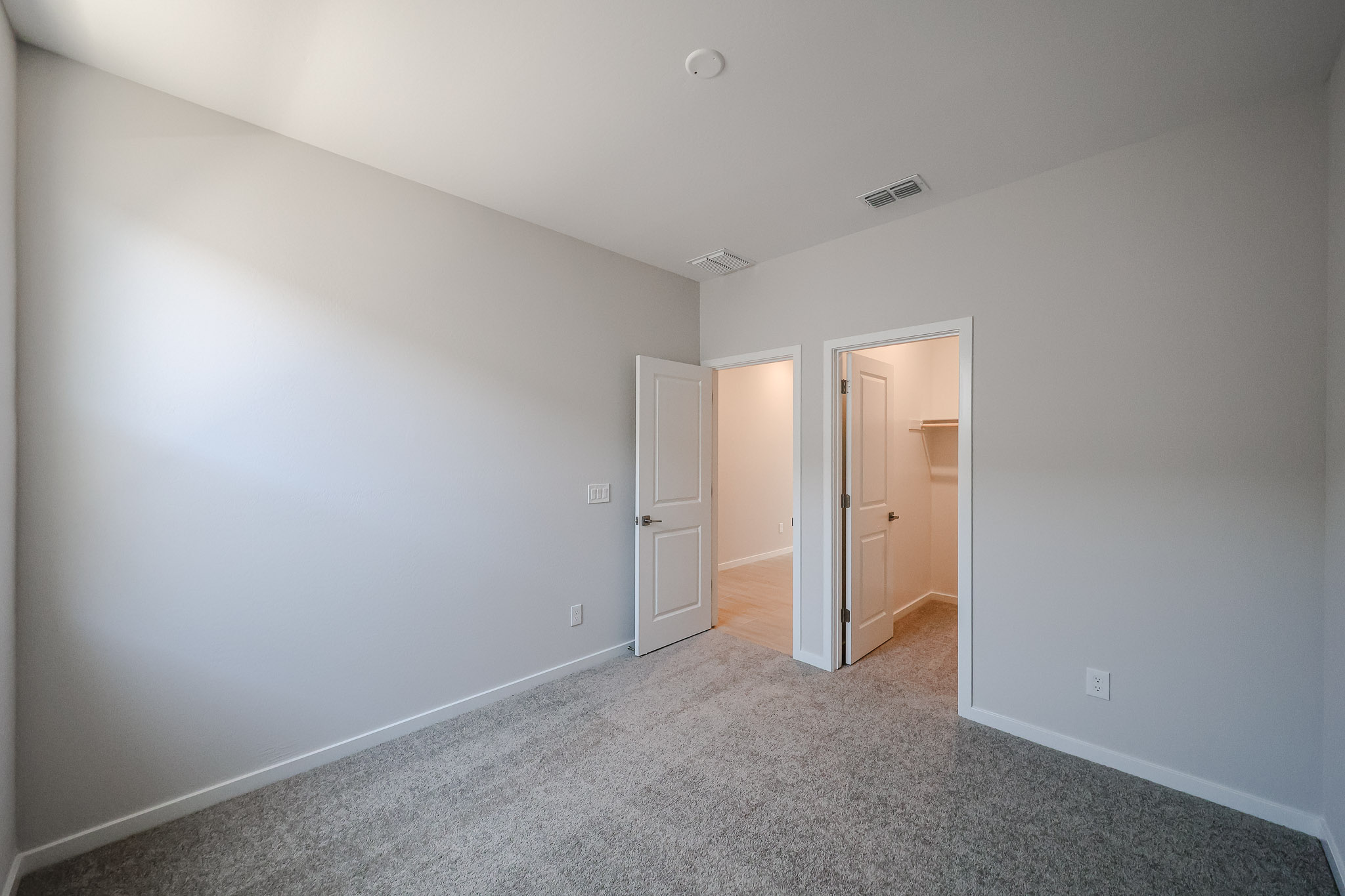 Empty room with gray carpet, white walls, and open doors leading to a hallway and a walk-in closet. Ceiling vent and recessed light are visible.