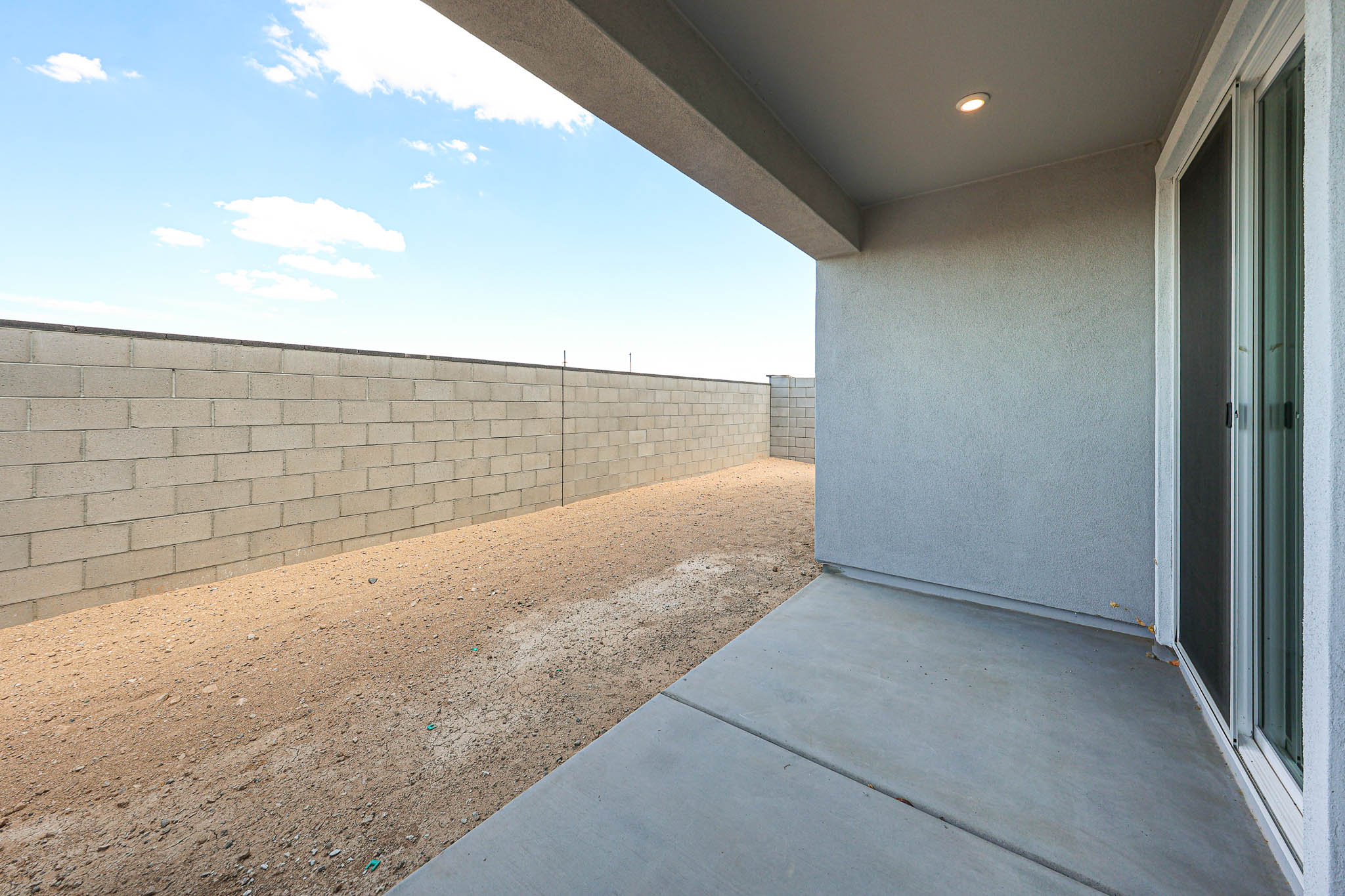 Covered concrete patio with a sliding glass door opens to a bare, fenced backyard featuring a tall cinder block wall under a blue sky with some clouds.