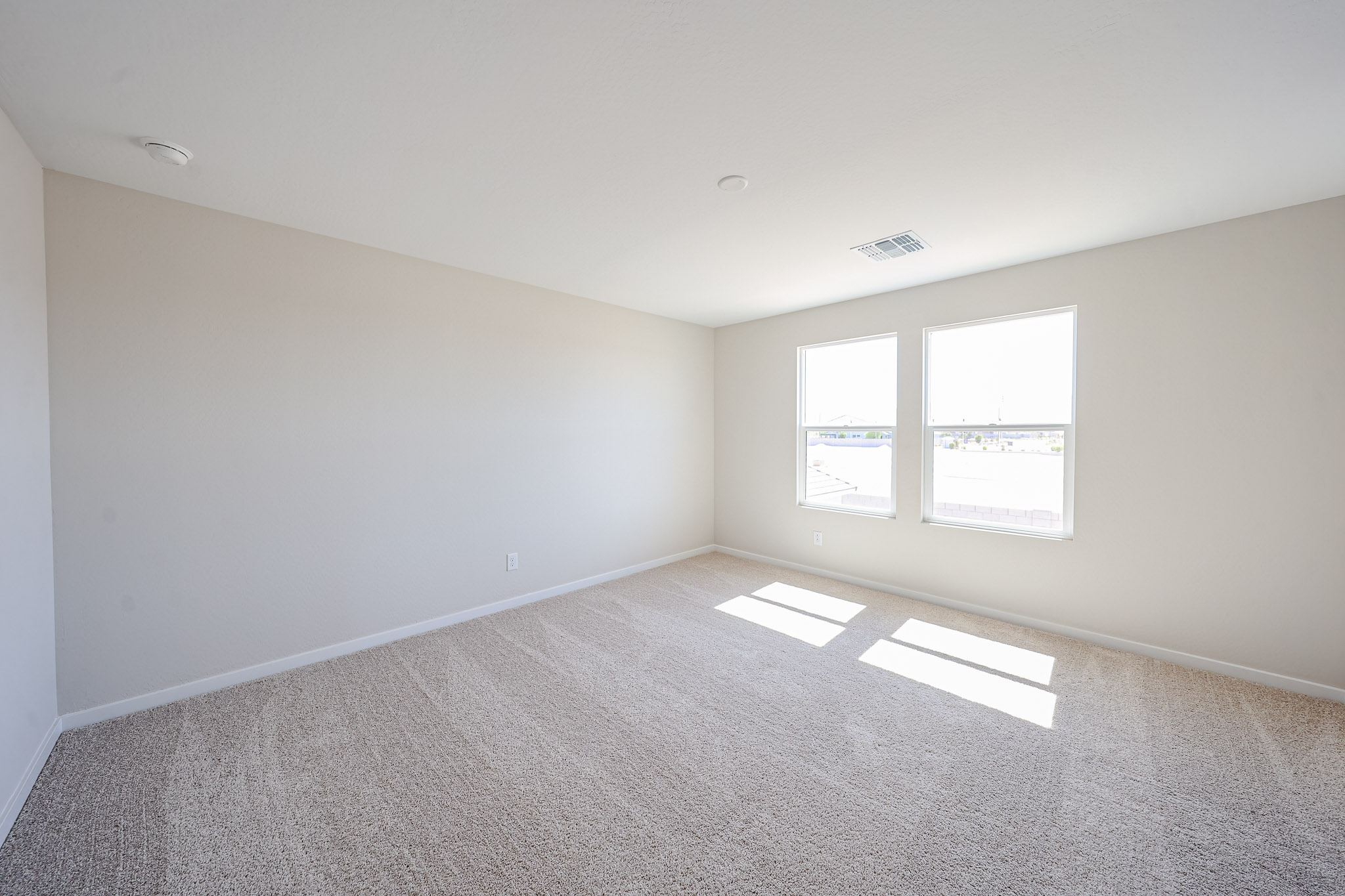 Empty, carpeted room with off-white walls and two large windows allowing natural light to enter. Ceiling vent and smoke detector are visible. Room appears clean and spacious.