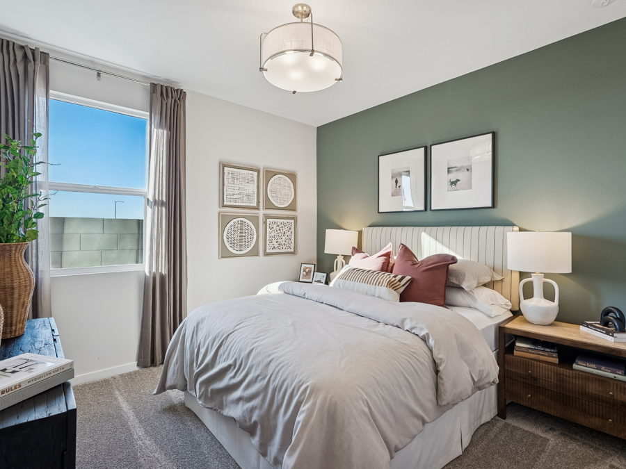 Bedroom with a green accent wall, neatly made bed, two bedside tables with lamps, framed art, and a window with curtains letting in natural light.