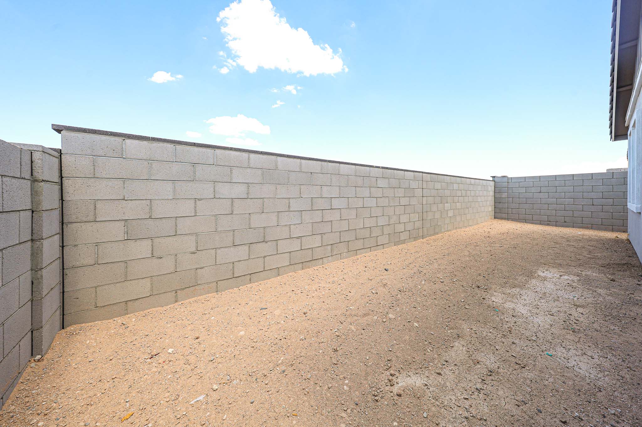 Enclosed backyard with a tall cinder block wall, bare dirt ground, and part of a gray building on the right under a blue sky with some clouds.
