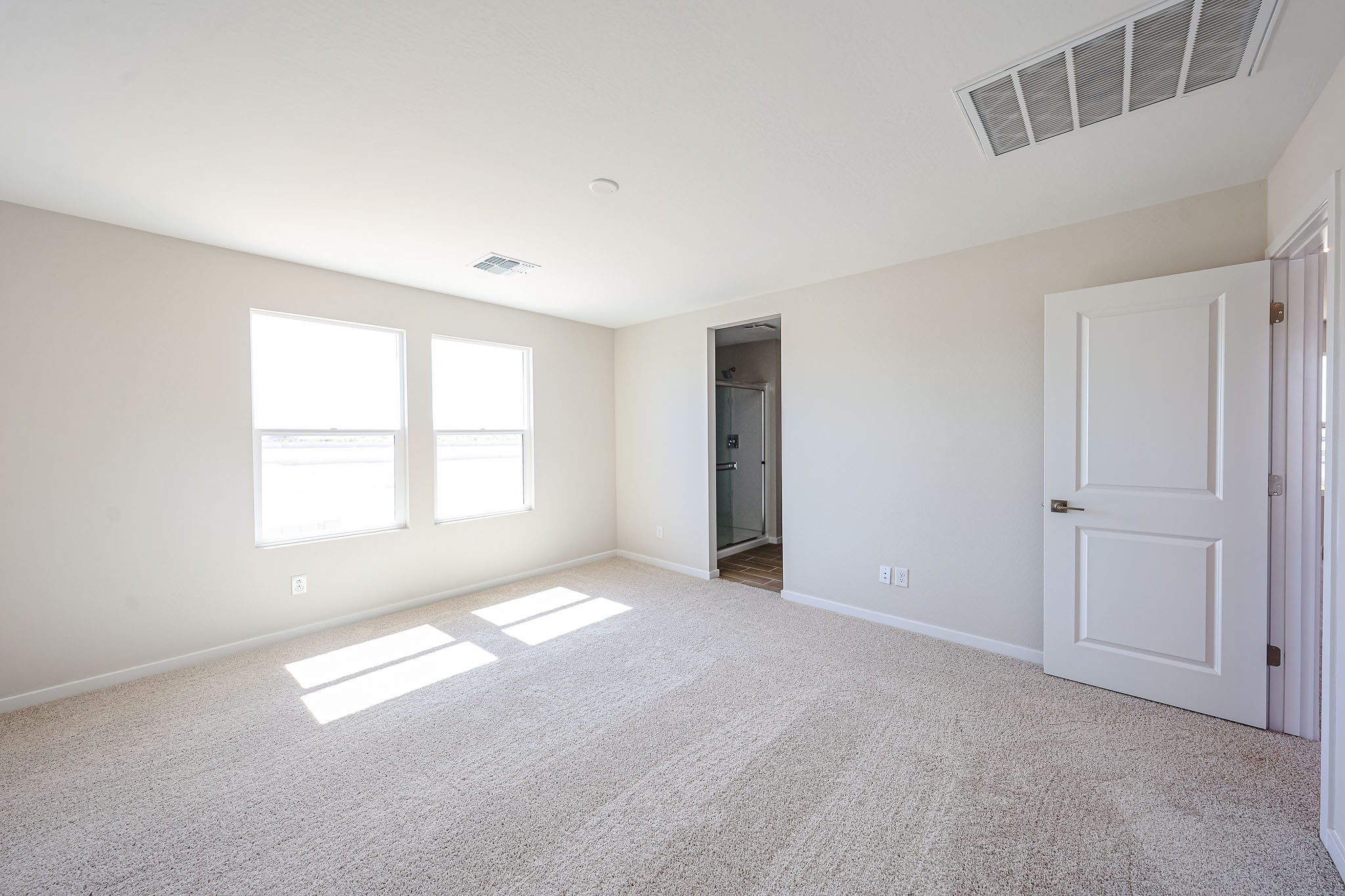 Empty, carpeted bedroom with two windows, white walls, and an open door leading to a bathroom. Sunlight streams through the windows onto the floor.