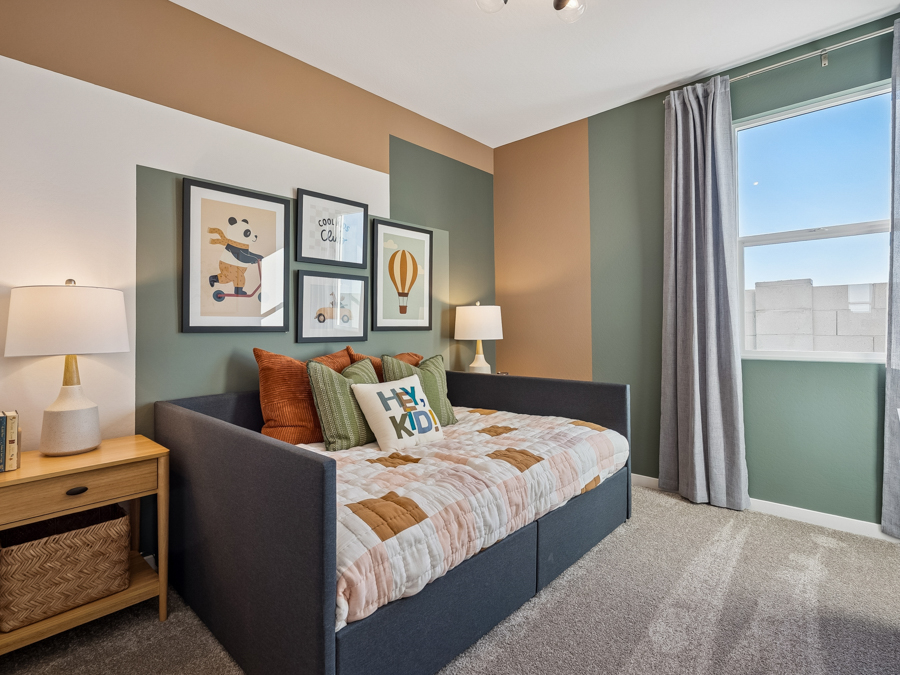 Children’s bedroom with a dark daybed, colorful pillows, framed animal and balloon art, two lamps on wooden tables, and a window with gray curtains letting in natural light.