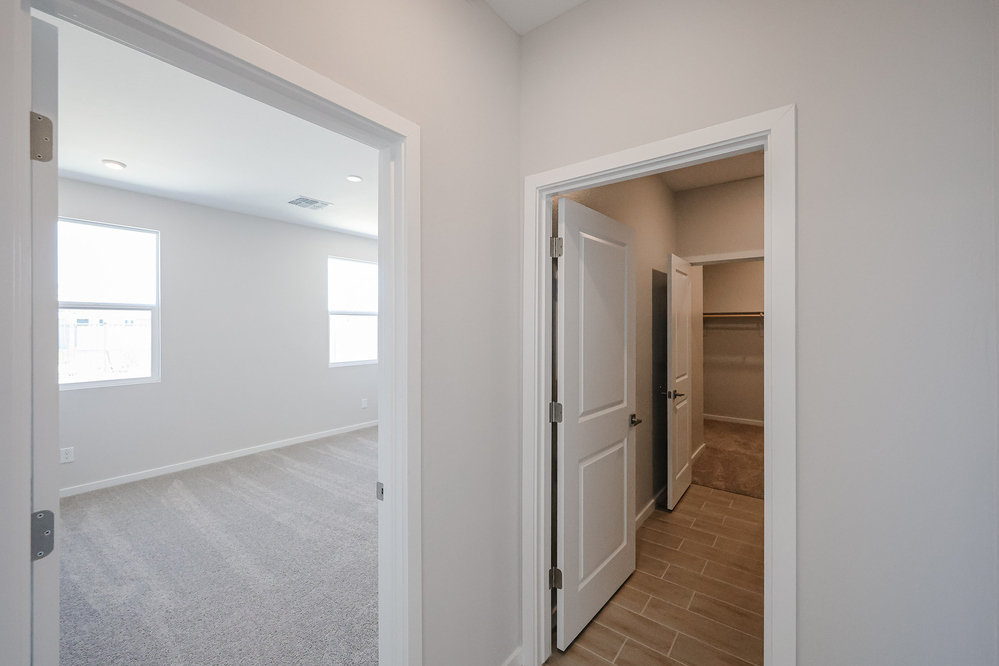 View of a hallway with two open doors, one leading to a carpeted bedroom with windows, and the other to a tiled walk-in closet or storage space.