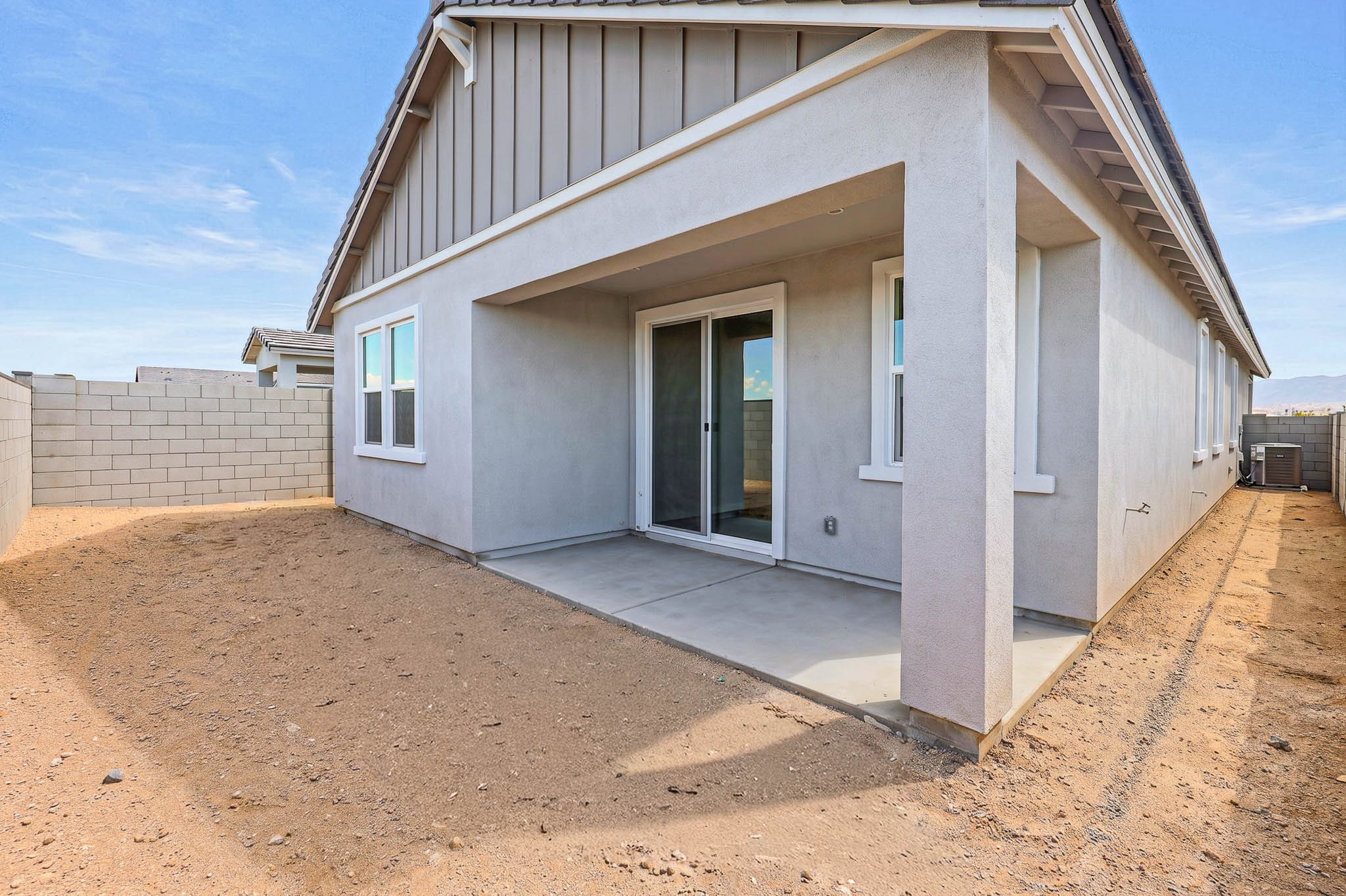 Backyard view of a modern house with a covered patio, sliding glass doors, and a bare dirt yard enclosed by a cinder block wall.