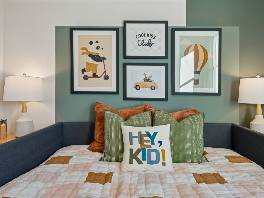 A neatly made bed with colorful pillows is set against a green accent wall, featuring four framed children’s prints and two table lamps on either side.
