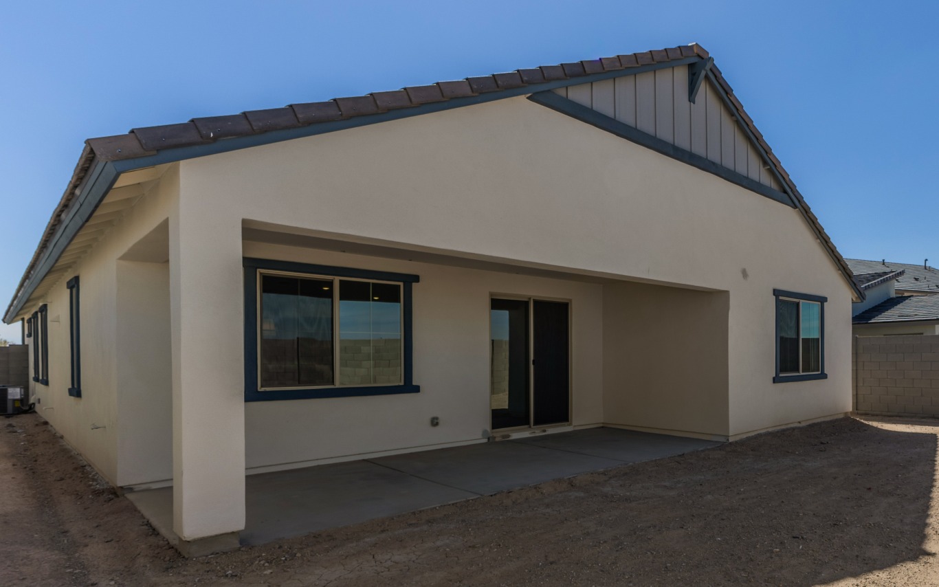 Exterior view of the back of a single-story house featuring a covered patio, sliding glass doors, and a dirt yard.