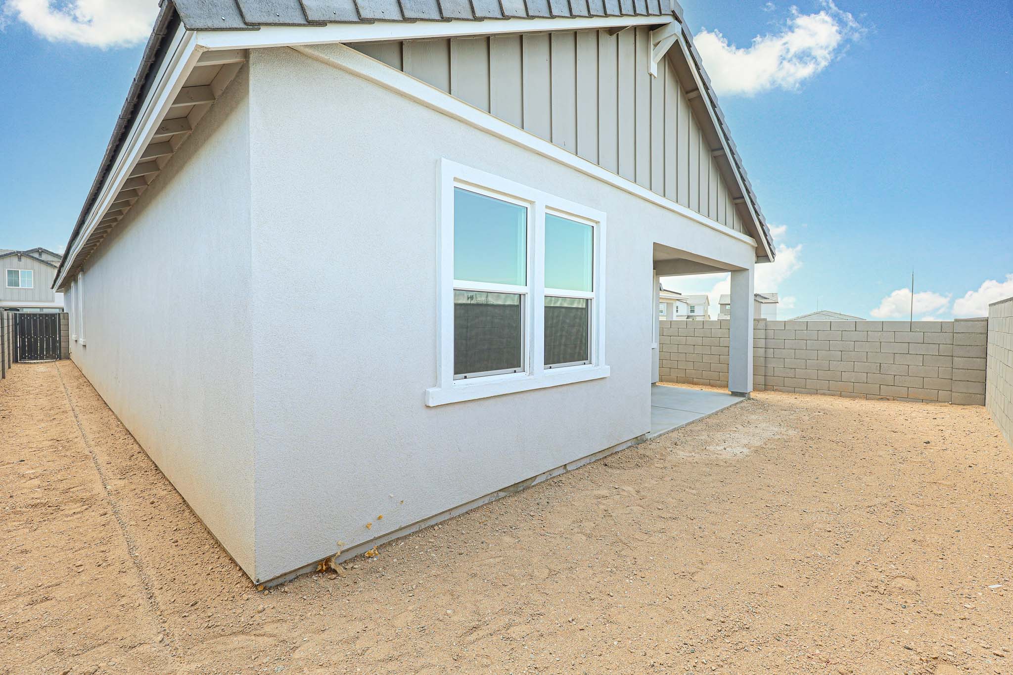 A side view of a modern house with white walls, two windows, a small covered patio, and a fenced backyard with bare dirt ground.