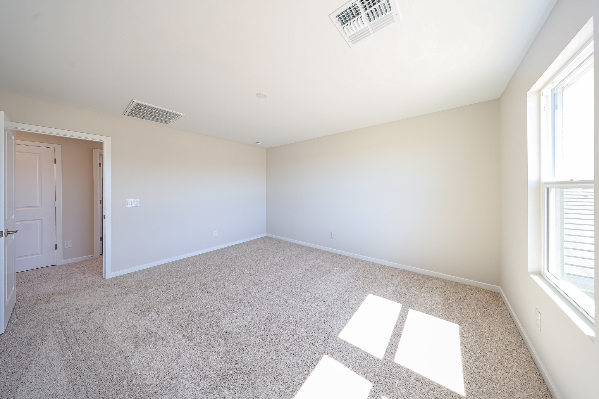 Empty room with beige carpet, white walls, an open door, and a window letting in natural light. Ceiling vents are visible.