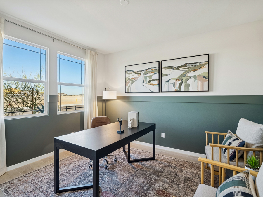 Modern home office with a black desk, office chair, armchair, abstract wall art, two windows, and a patterned rug.