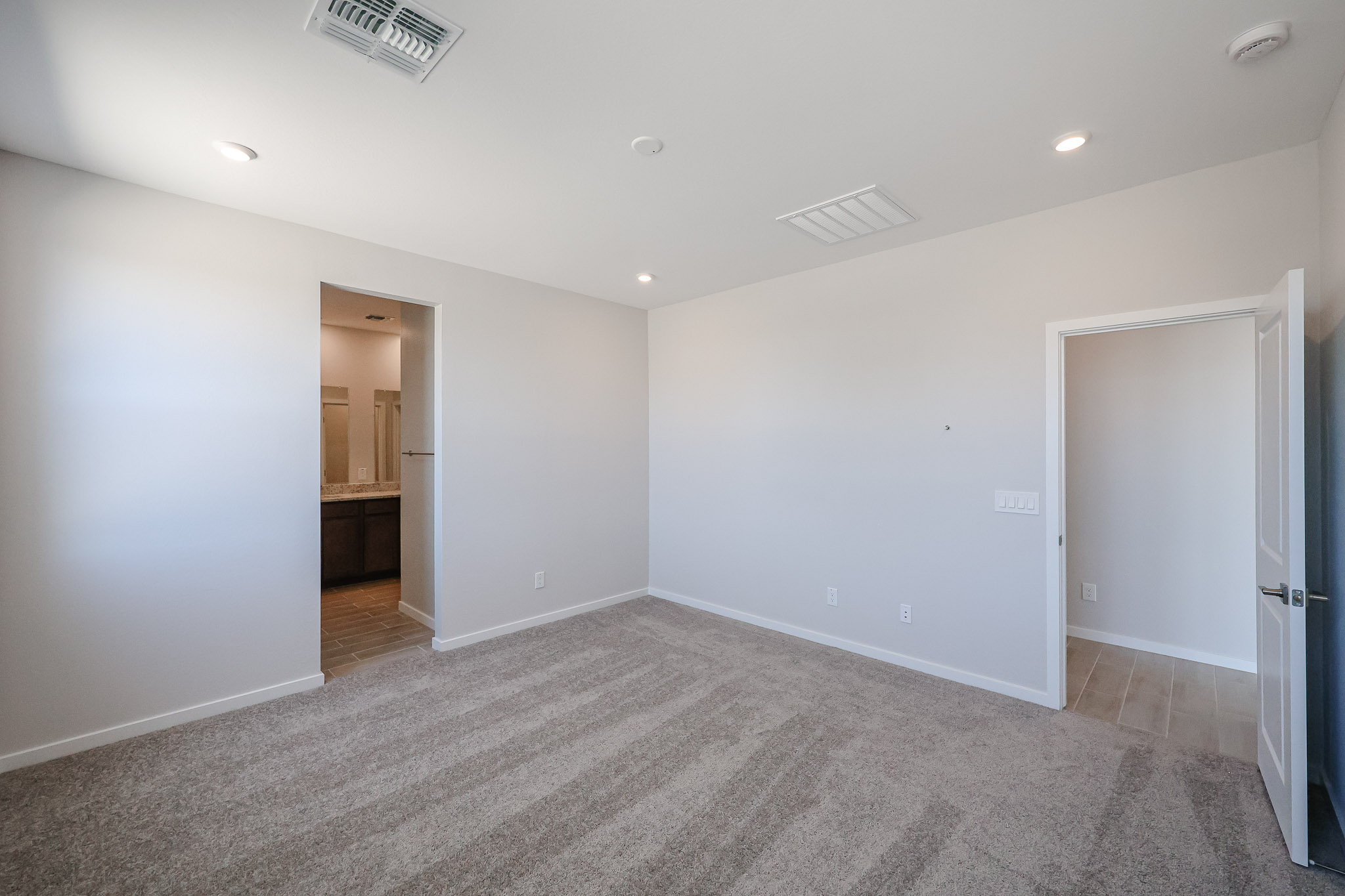 Empty bedroom with beige carpet, white walls, recessed ceiling lights, and two doorways leading to a bathroom and hallway.