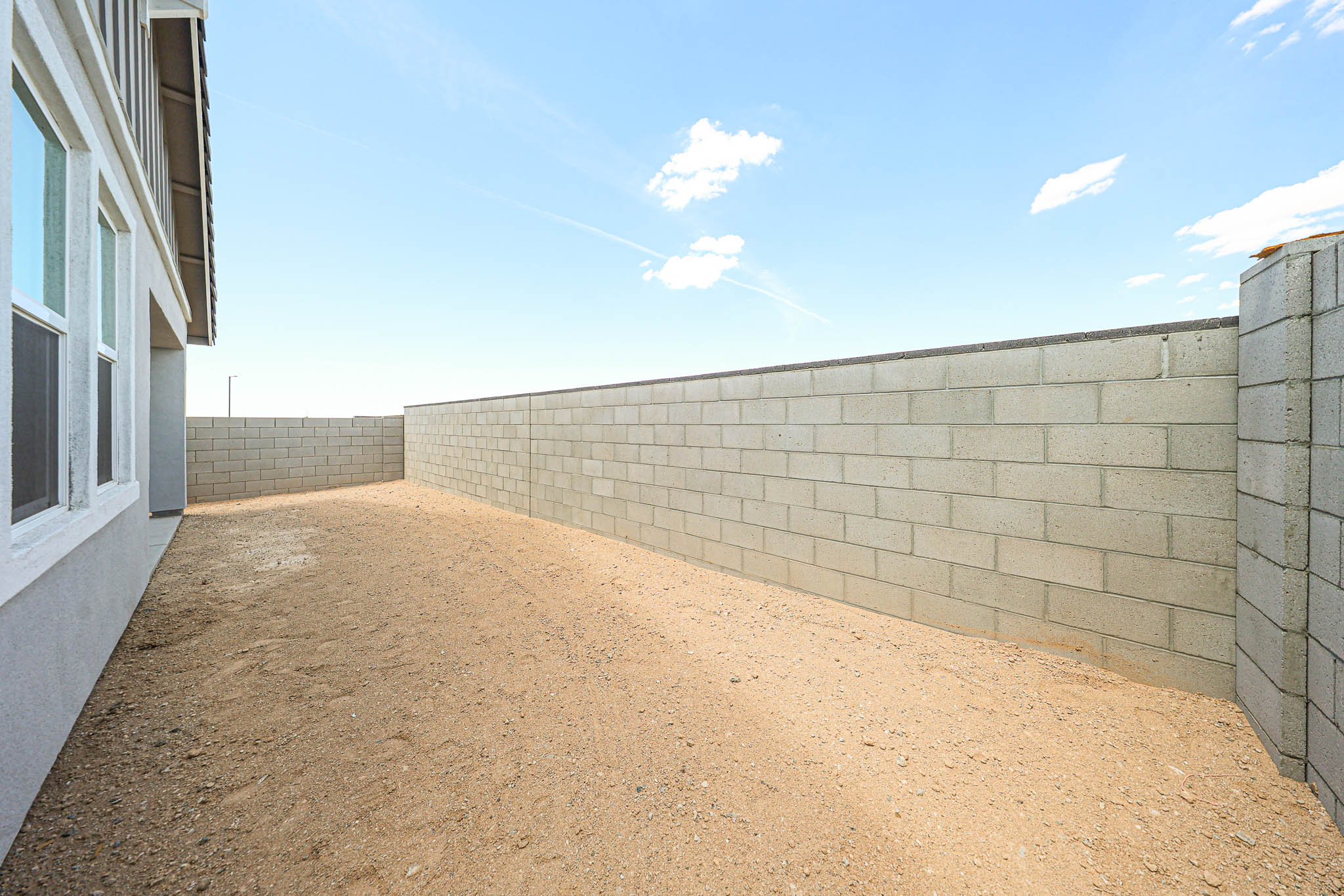 A sandy backyard enclosed by a tall, gray concrete block wall, with part of a white house visible on the left under a clear blue sky.