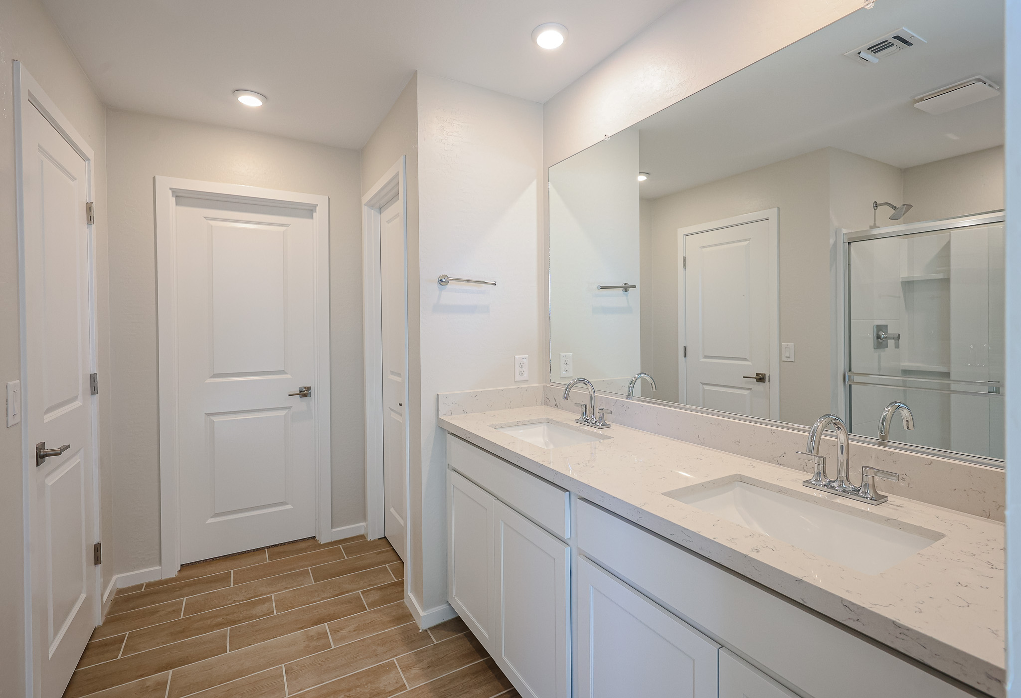 Modern bathroom with double sinks, quartz countertop, white cabinets, large mirror, wood-look tile floor, two doors, and a glass shower enclosure in the background.