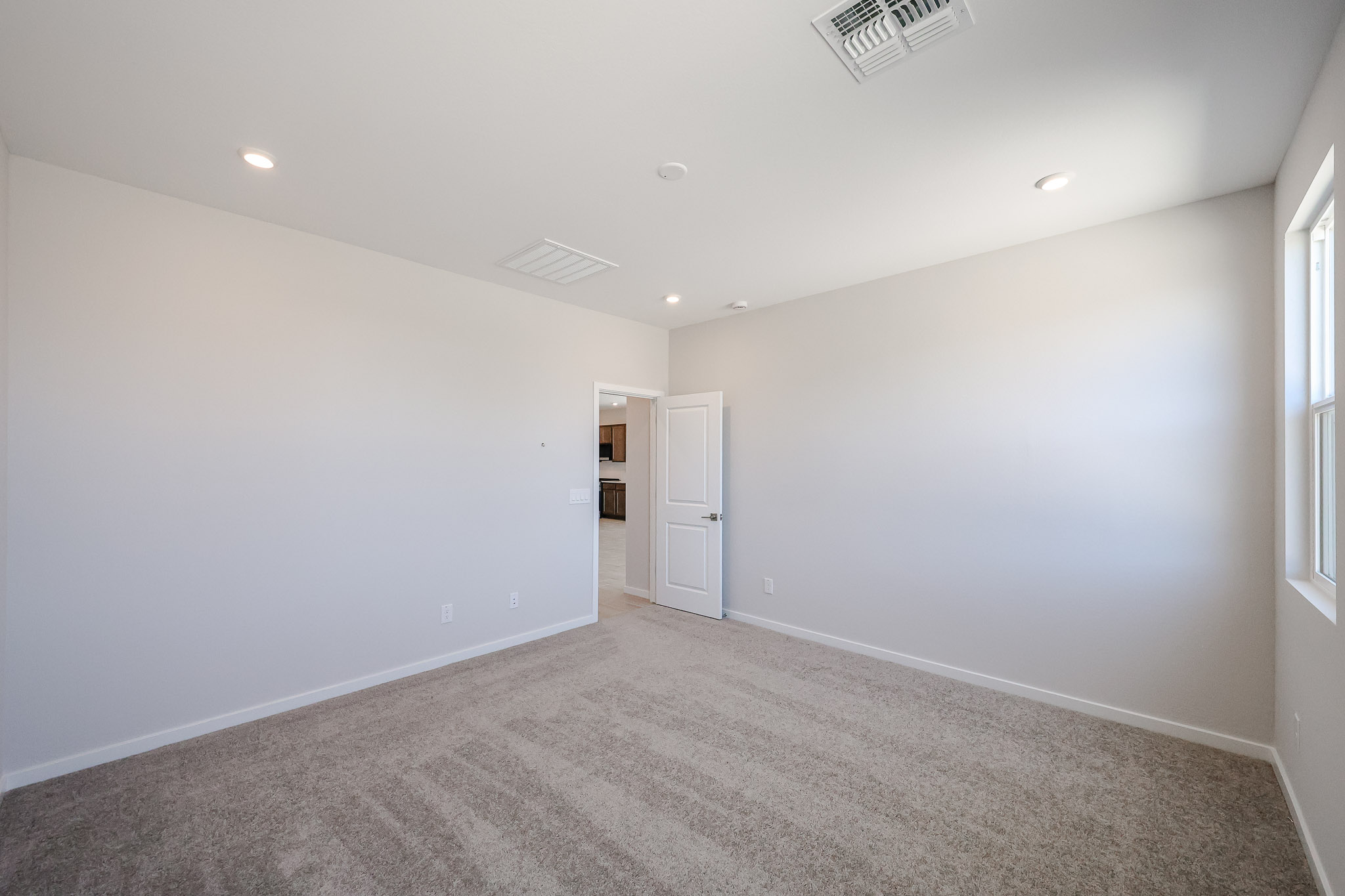 Empty carpeted room with white walls, ceiling lights, a window on the right, and an open door leading to another area of the house.