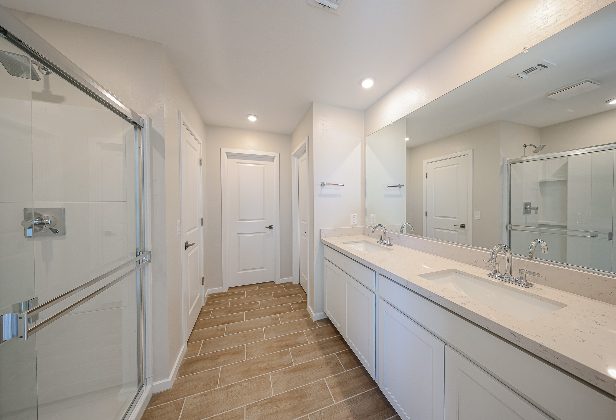 Modern bathroom with dual sinks, large mirror, glass shower enclosure, white cabinets, beige tile floor, and recessed lighting.
