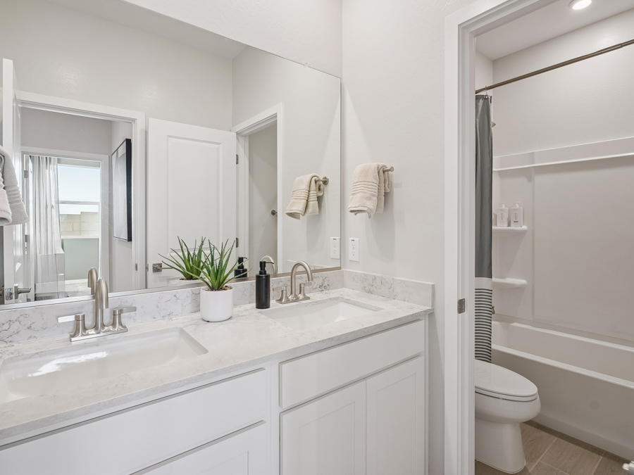 Modern bathroom with double sinks, white cabinets, a marble countertop, a potted plant, and a view of a shower and toilet reflected in a large mirror.