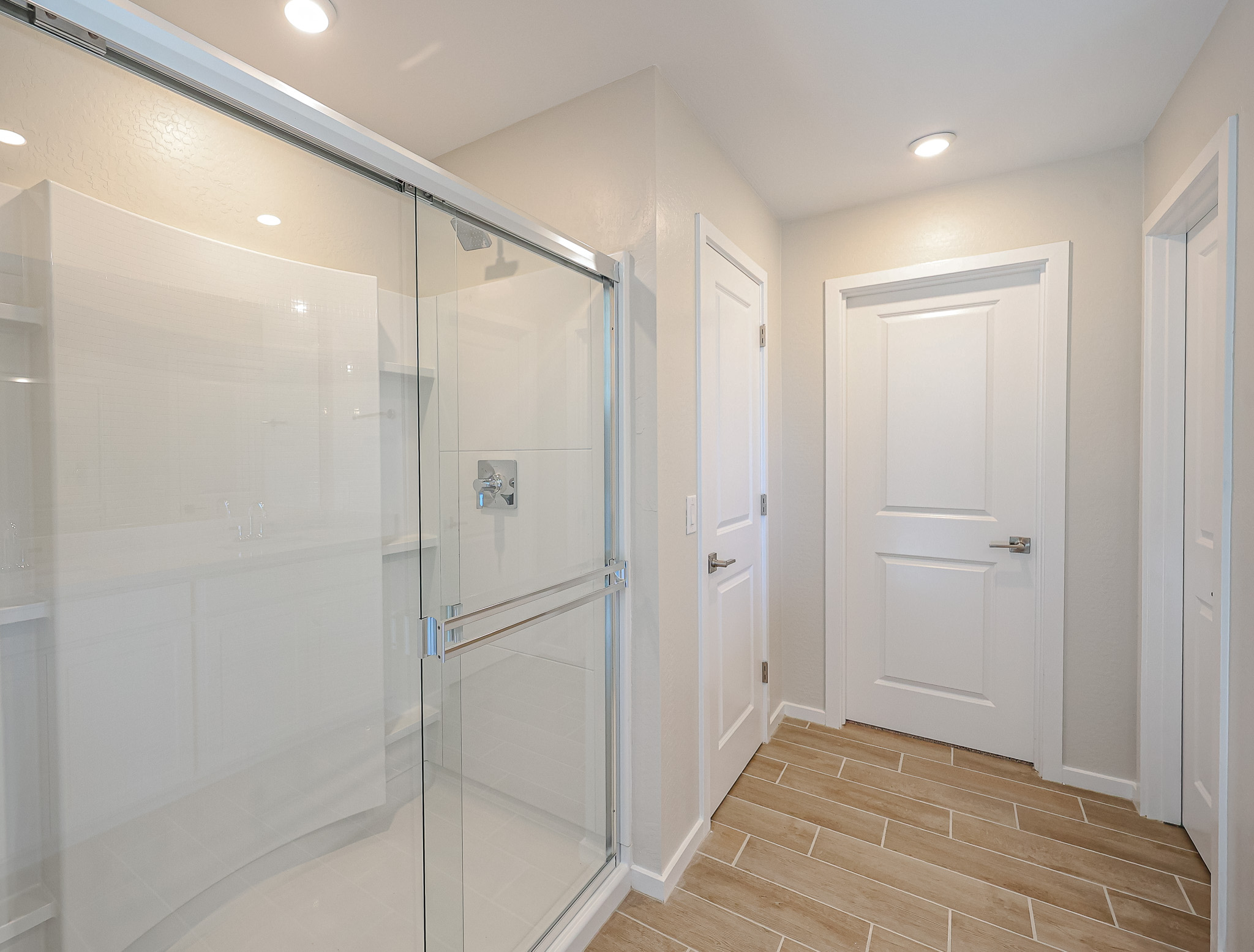 Modern bathroom with wood-look tile flooring, a glass-enclosed shower, white walls, recessed lighting, and two closed white doors.